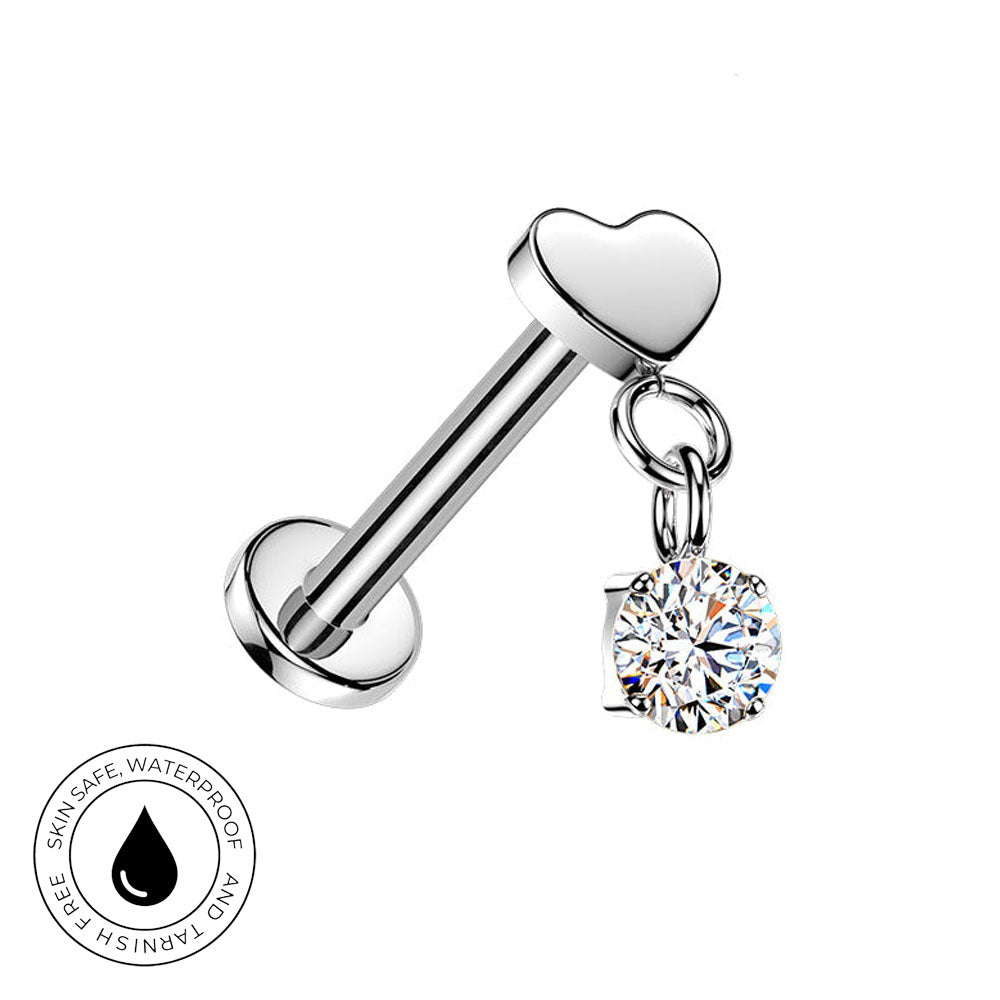 A silver-toned barbell-style body jewelry piece with a heart-shaped end and a dangling clear round gemstone charm, shown on a white background with a small skin-safe waterproof badge in the lower left.