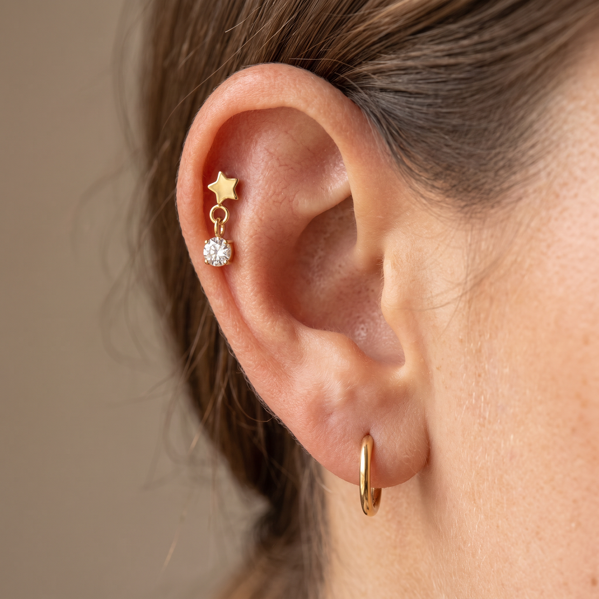 A close-up of a person's ear showcasing a gold star and clear crystal stud earring on the upper ear cartilage, alongside a simple gold hoop earring in the lobe.