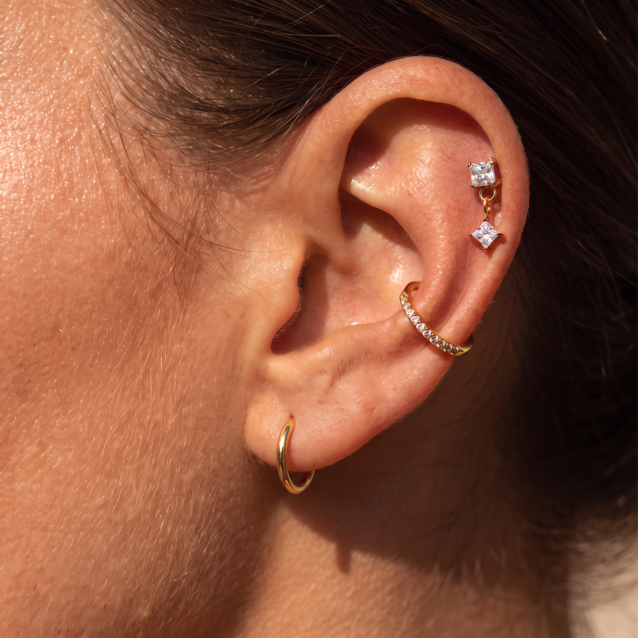 Close-up of a person's ear showcasing multiple gold and silver stud earrings, including a small gold hoop, a gold ear cuff with clear stones, and a double-drop silver stud with a clear star-shaped gem.