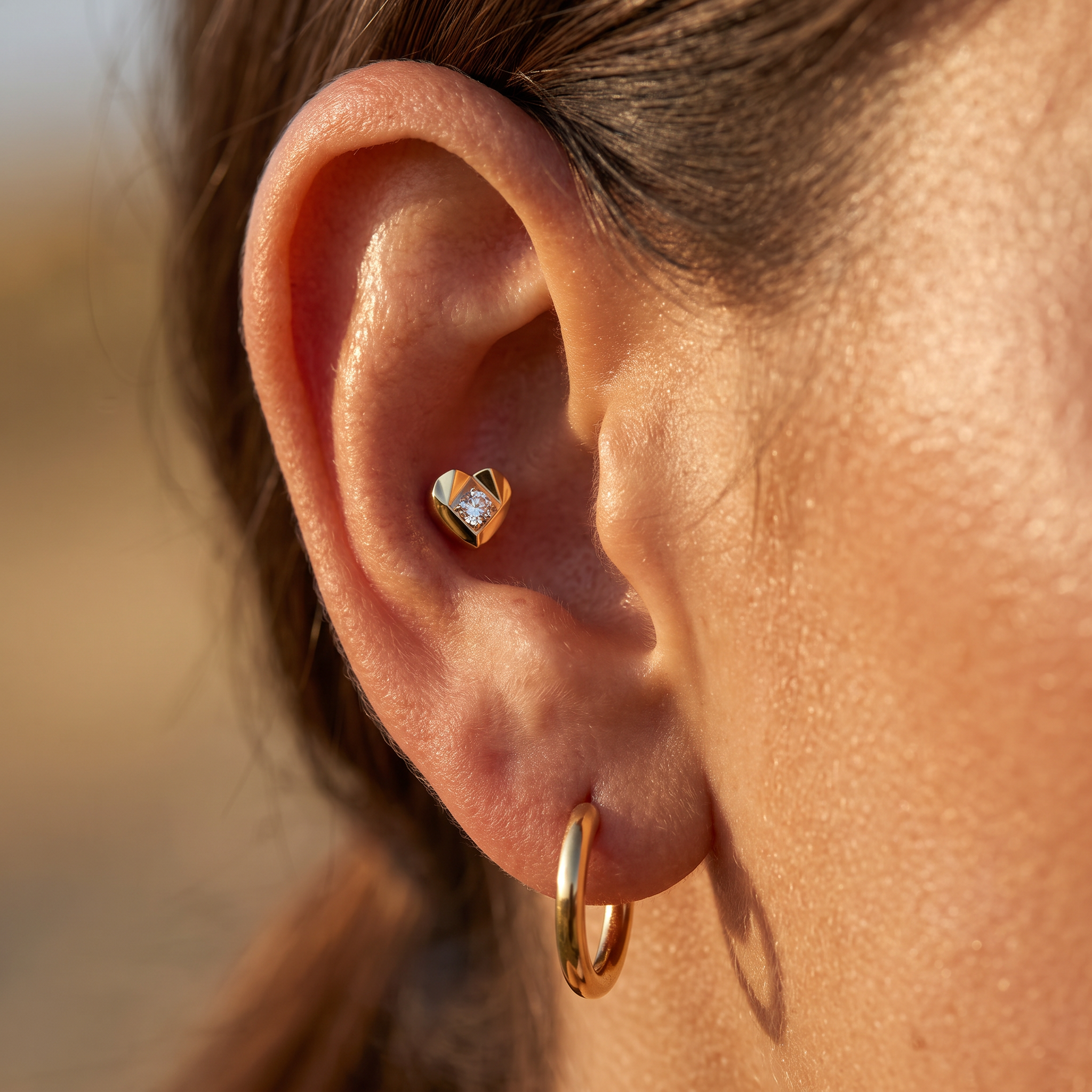 A close-up of a person's ear showcasing two gold earrings: a small heart-shaped stud earring with a clear gemstone and a classic gold hoop earring.