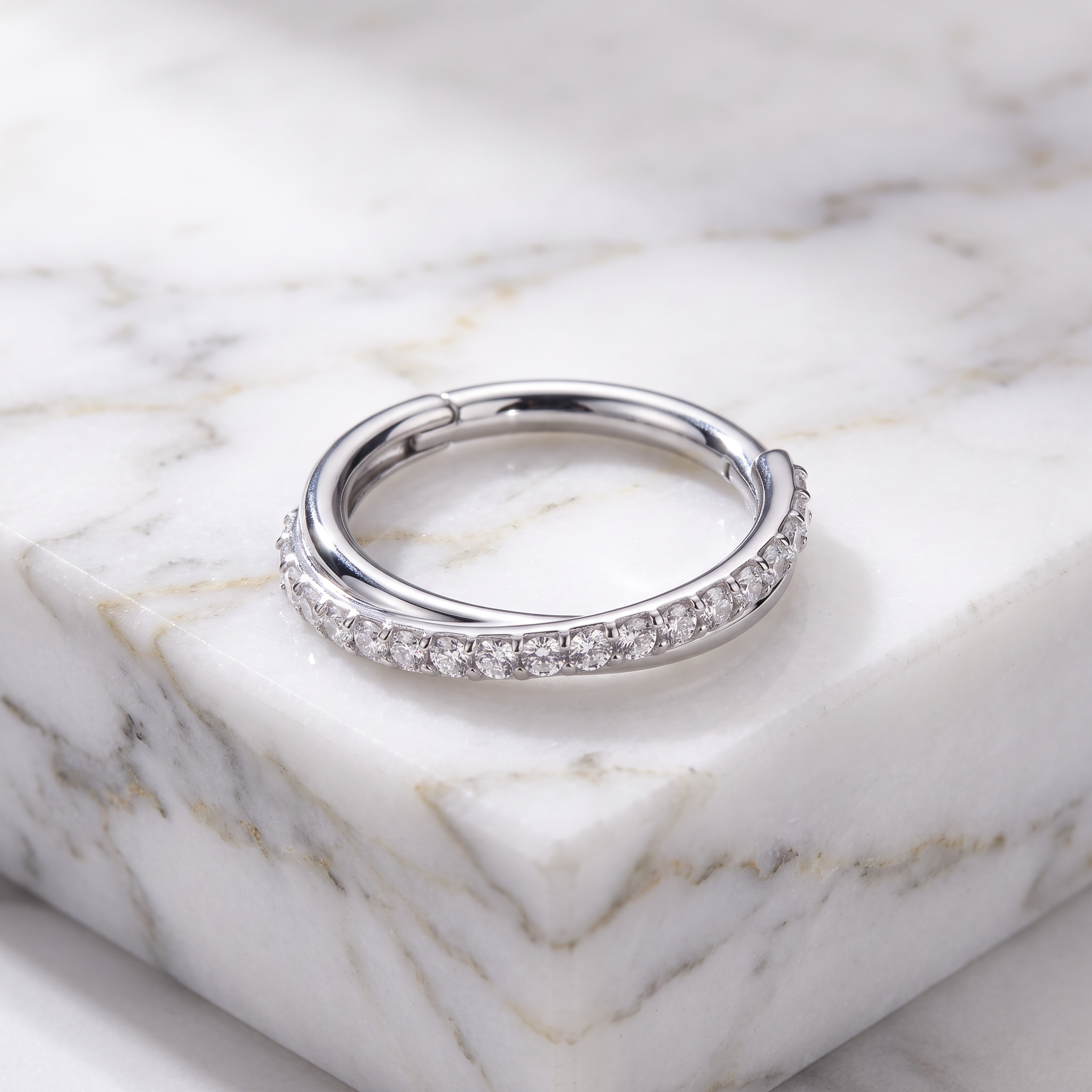 A silver-toned huggie ring with a row of clear pavé stones on one side, resting on a white marble block.
