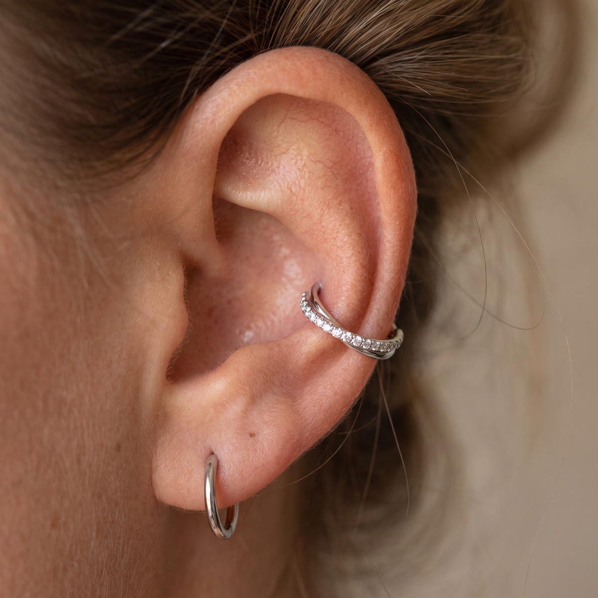 A close-up of a person's ear showcasing two silver piercings: a small hoop earring in the lobe and a curved ear cuff adorned with small clear stones, set against a soft beige background.