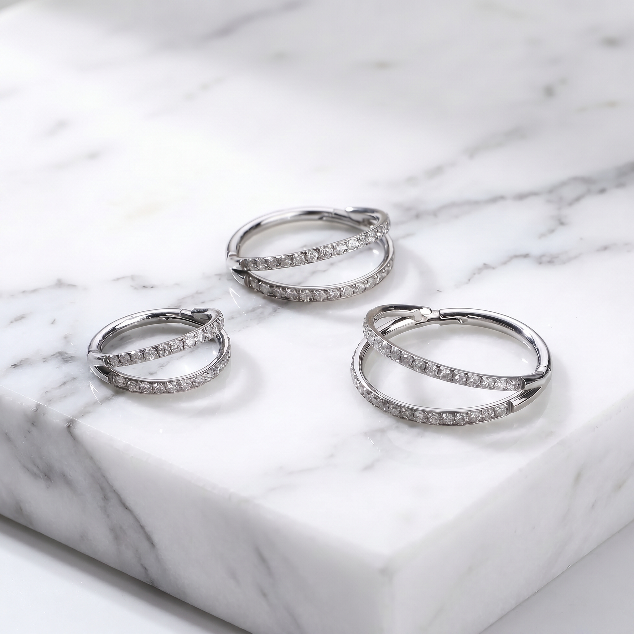 Three silver hoop earrings with pavé-set clear stones, arranged on a white marble slab.