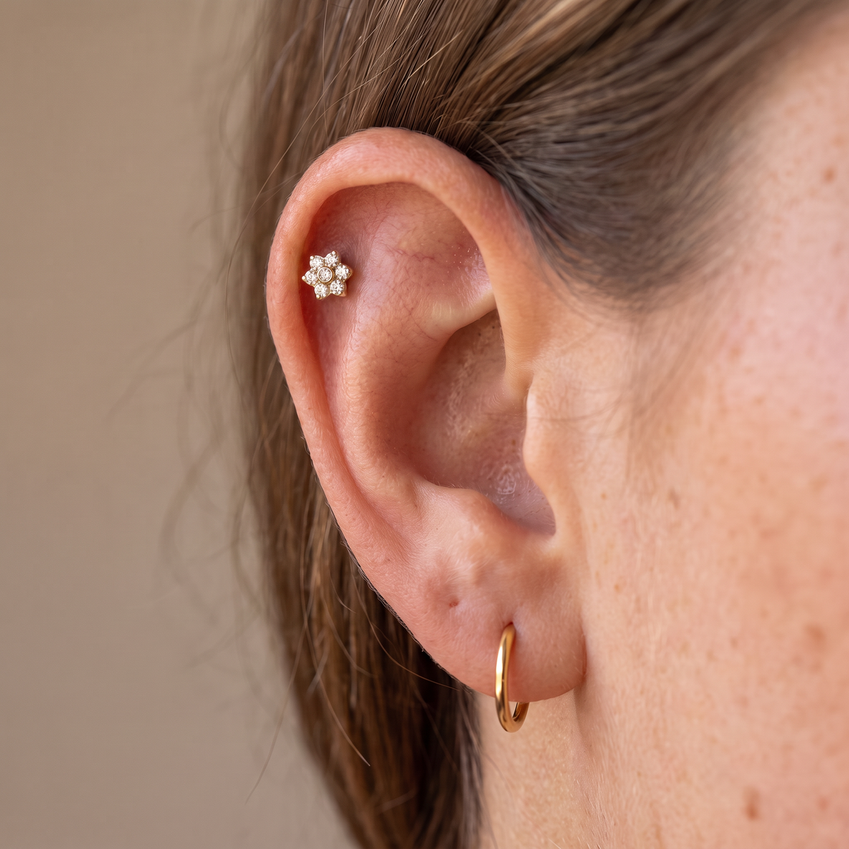 A close-up of a person's ear showcasing two small gold earrings: a delicate gold hoop in the lobe and a small gold flower-shaped stud in the helix.