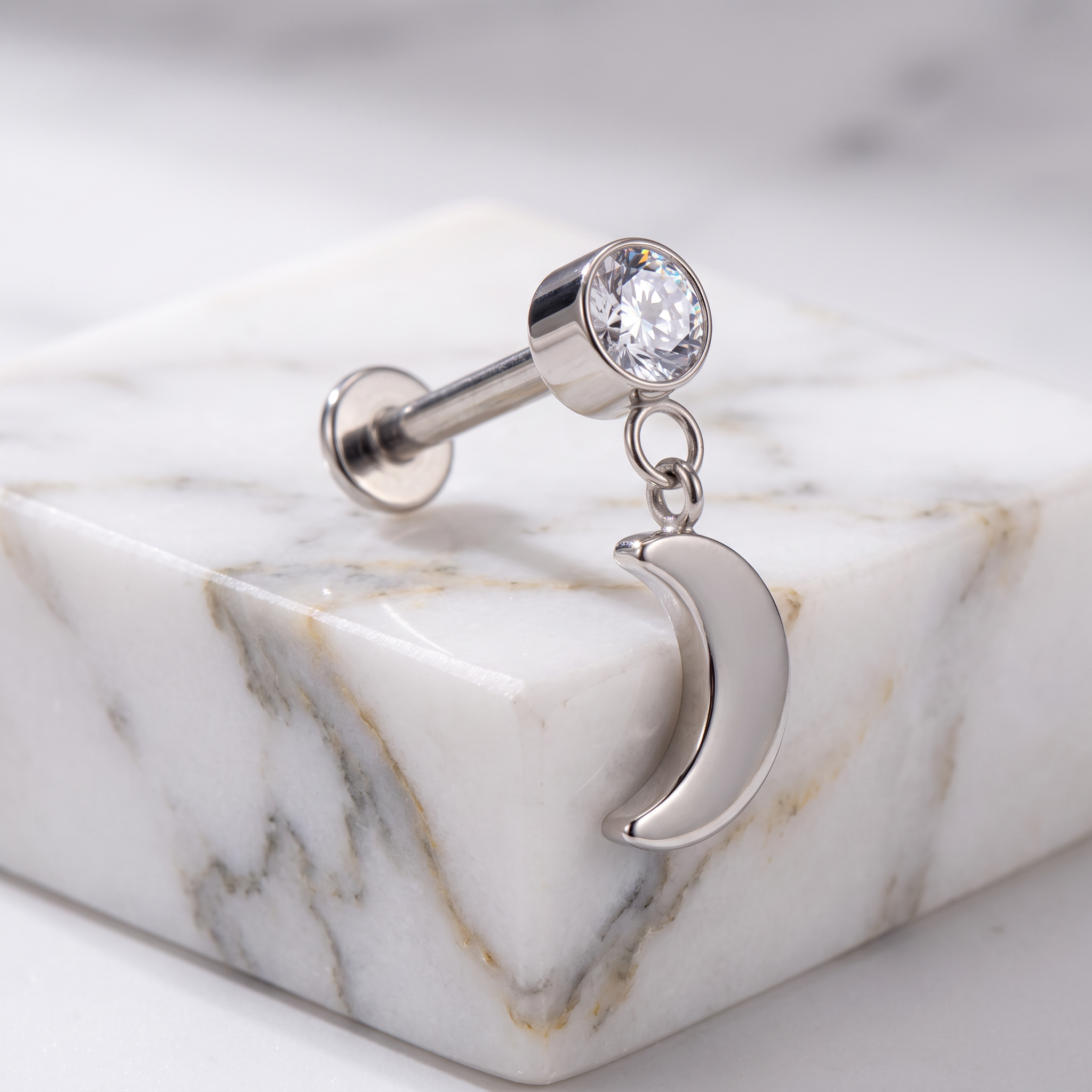 A silver-toned labret piercing stud with a clear round gemstone and a dangling crescent moon charm, displayed on a polished white marble block.