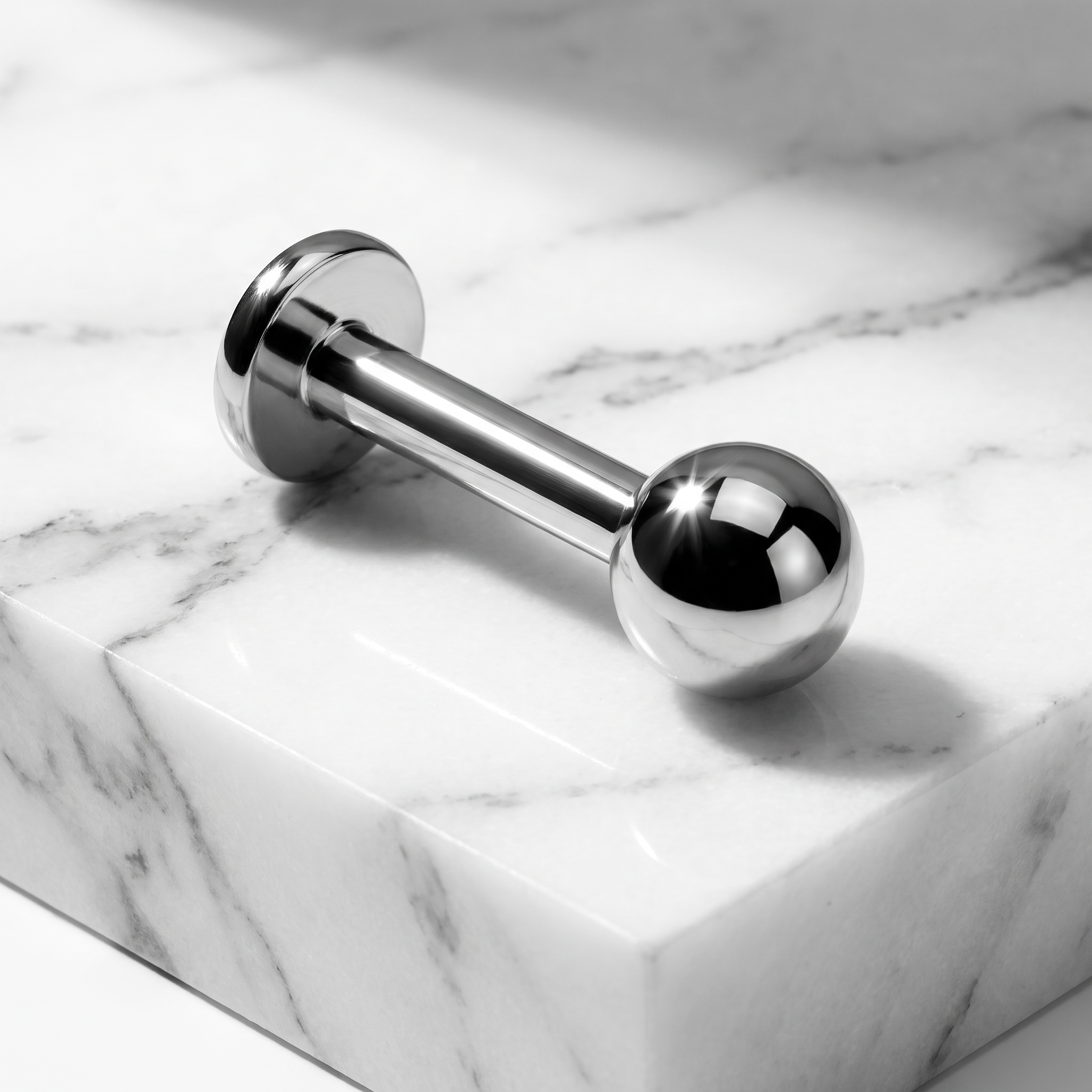 A polished stainless steel barbell piercing jewelry piece with spherical ends, resting diagonally on a white marble block with gray veining.