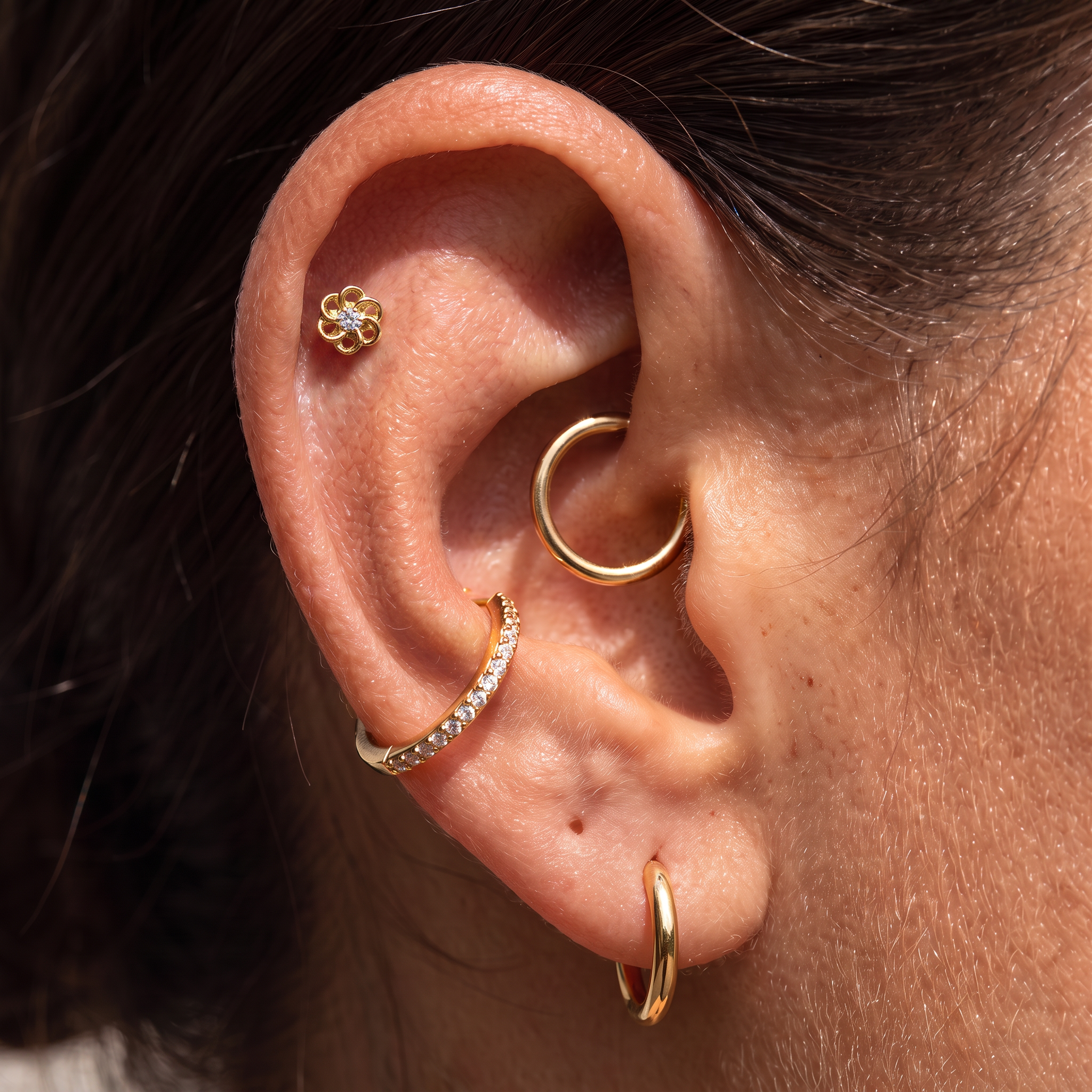 Close-up of a person's ear showcasing multiple gold ear piercings, including a small flower stud, a curved hoop, a diamond-accented huggie, and a lower hoop.