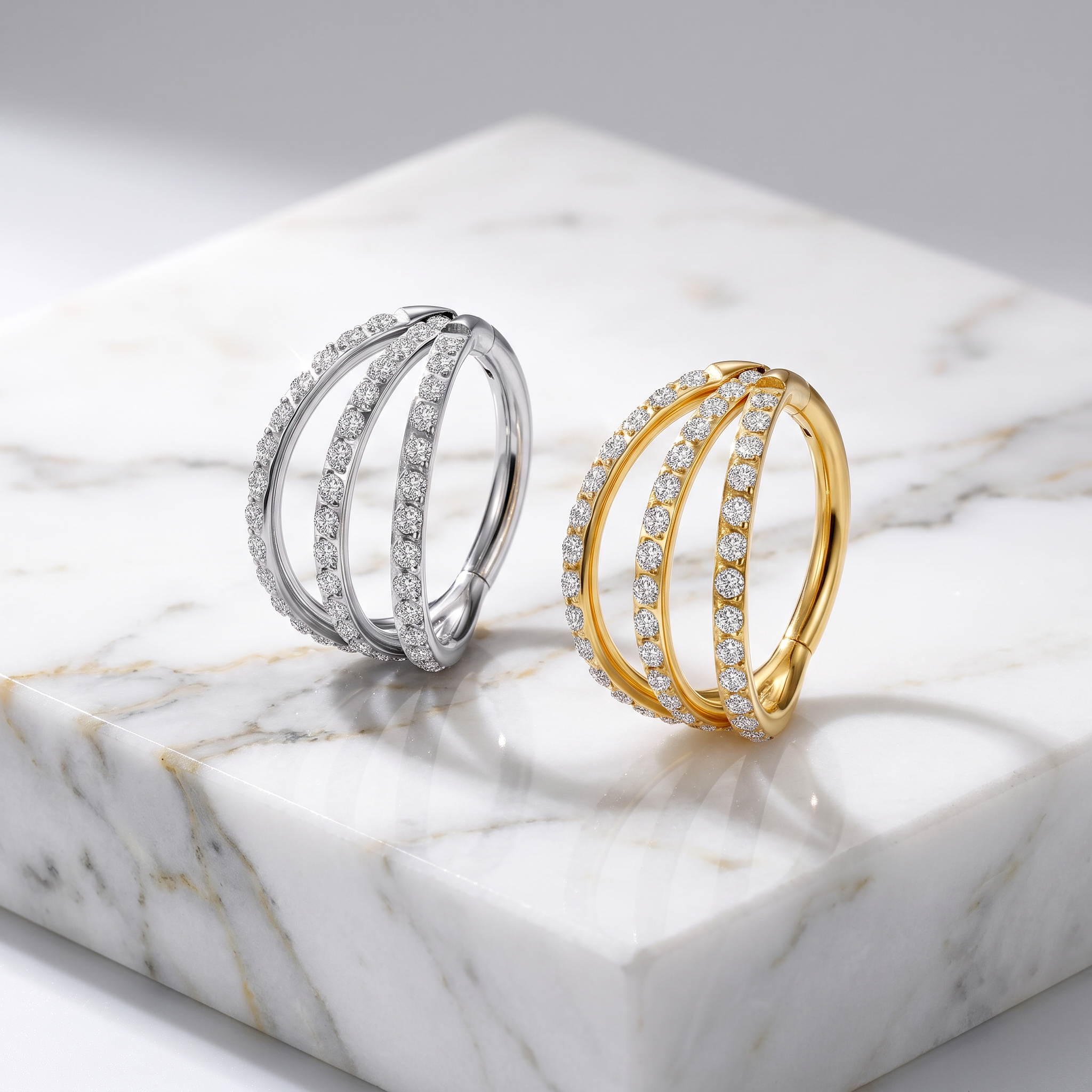 Two triple-band hoop rings with pavé-set stones, one in silver and one in yellow gold, displayed on a white marble block.