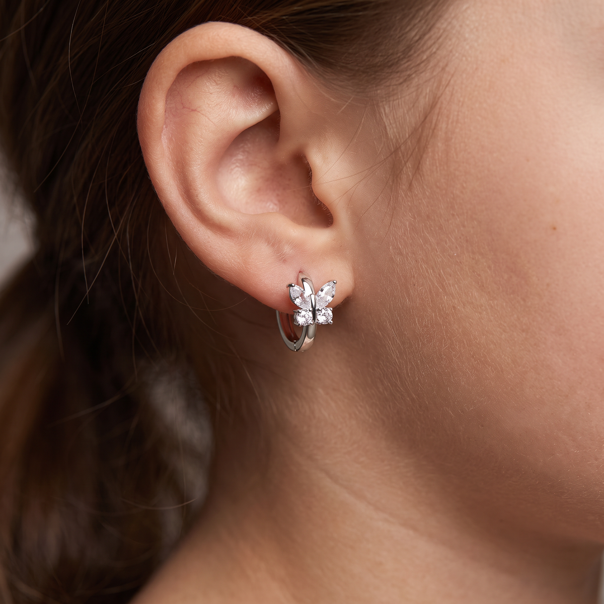 A close-up of a person's ear wearing a silver huggie hoop earring adorned with a small silver flower cluster set with clear stones.
