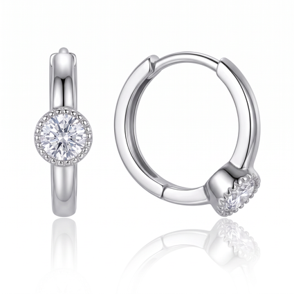 A pair of polished silver huggie hoop earrings, one shown from the front with a round clear gemstone in a halo setting and the other from the side showing the hinge and the gemstone setting, on a white background with a soft reflection.