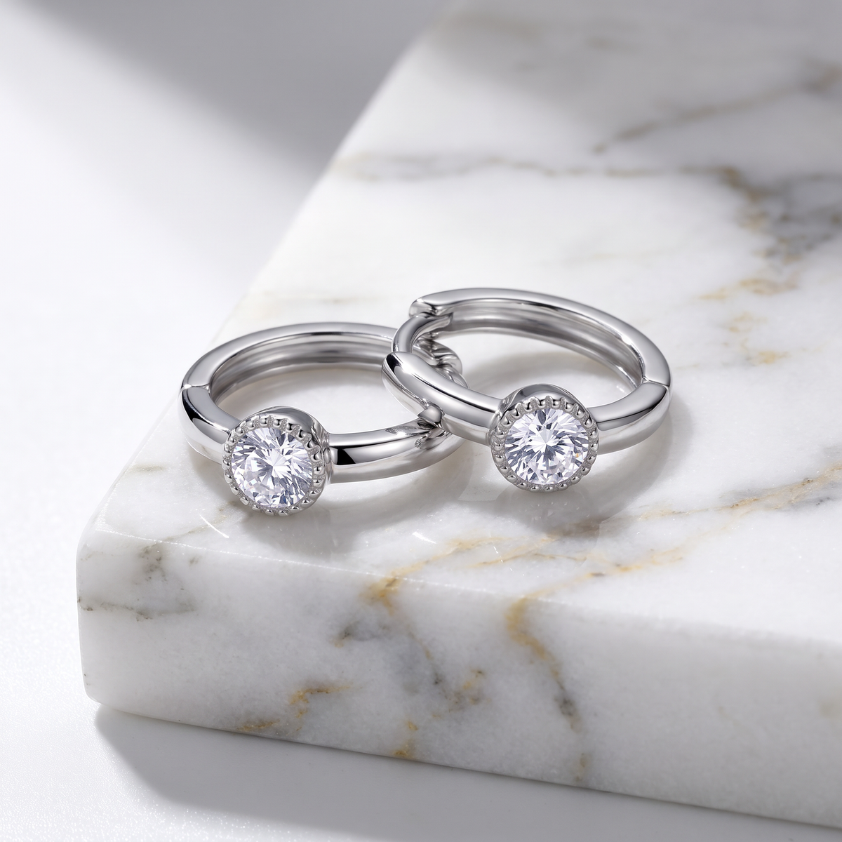 Two silver-toned huggie rings with round clear gemstones, displayed on a white marble block.
