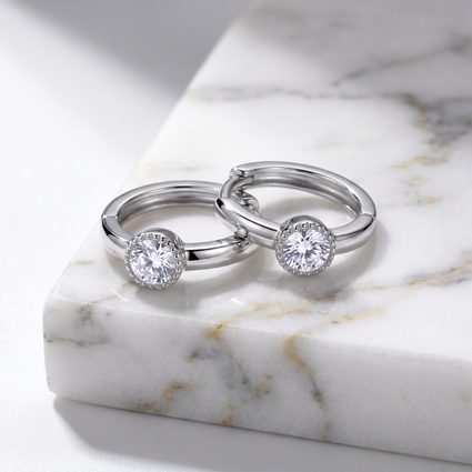 Two silver-toned huggie rings with round clear gemstones, displayed on a white marble block.