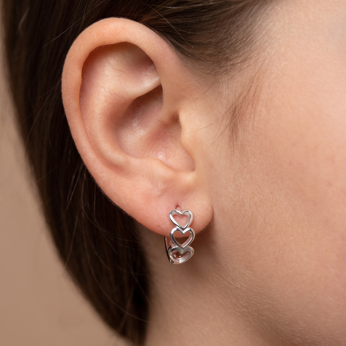 A close-up of a person's ear wearing a small silver hoop earring with two interlocking open heart shapes.