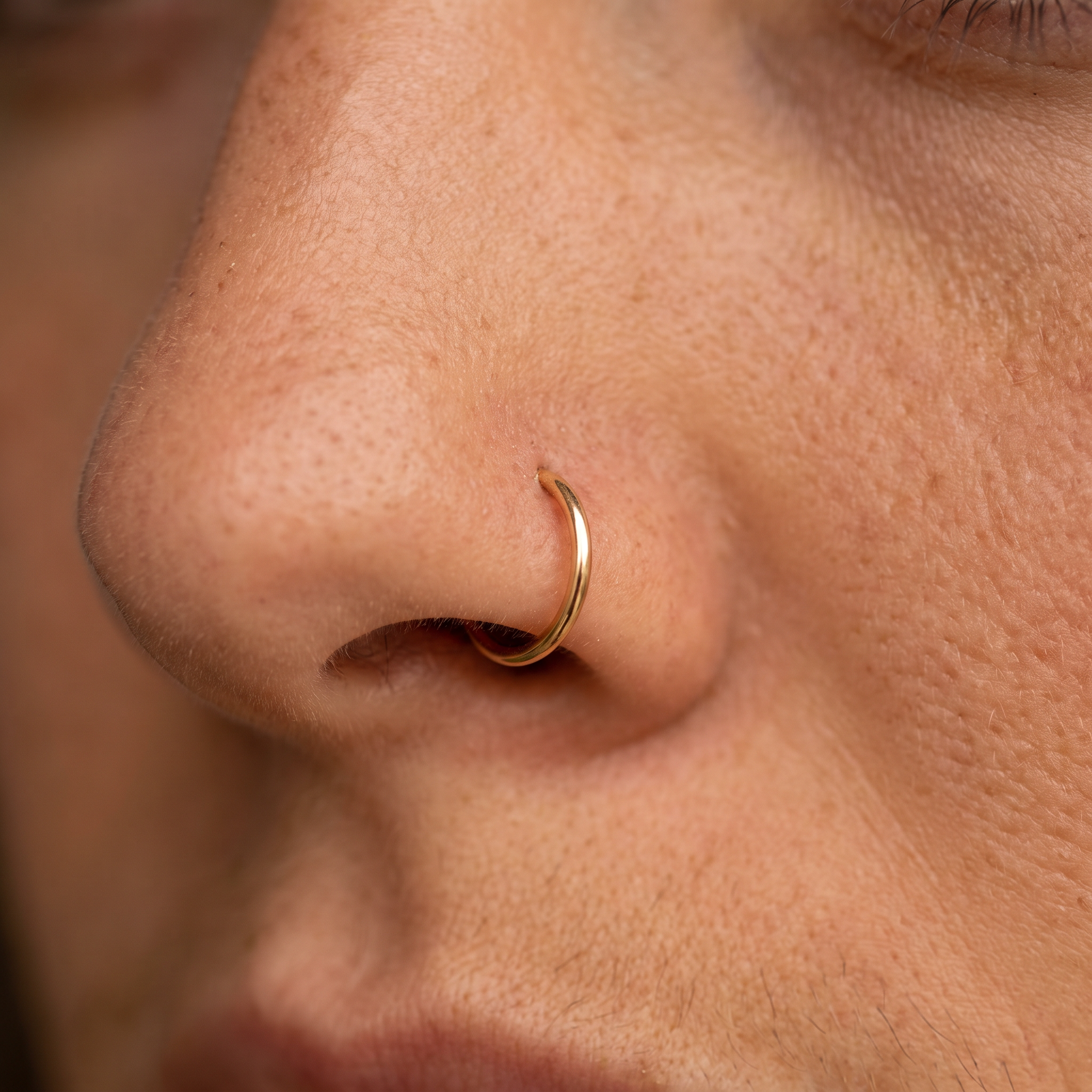 A close-up of a person's nose wearing a thin, polished gold nose hoop ring.