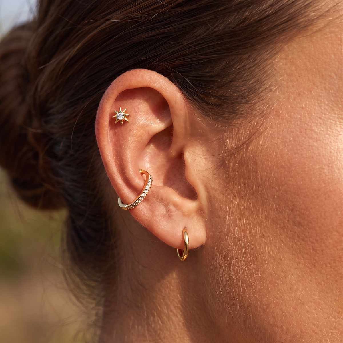 A close-up of a person's ear showcasing four gold ear piercings: a small starburst stud in the cartilage, a curved pavé hoop in the lobe, a small gold hoop in the earlobe, and a gold ear cuff.