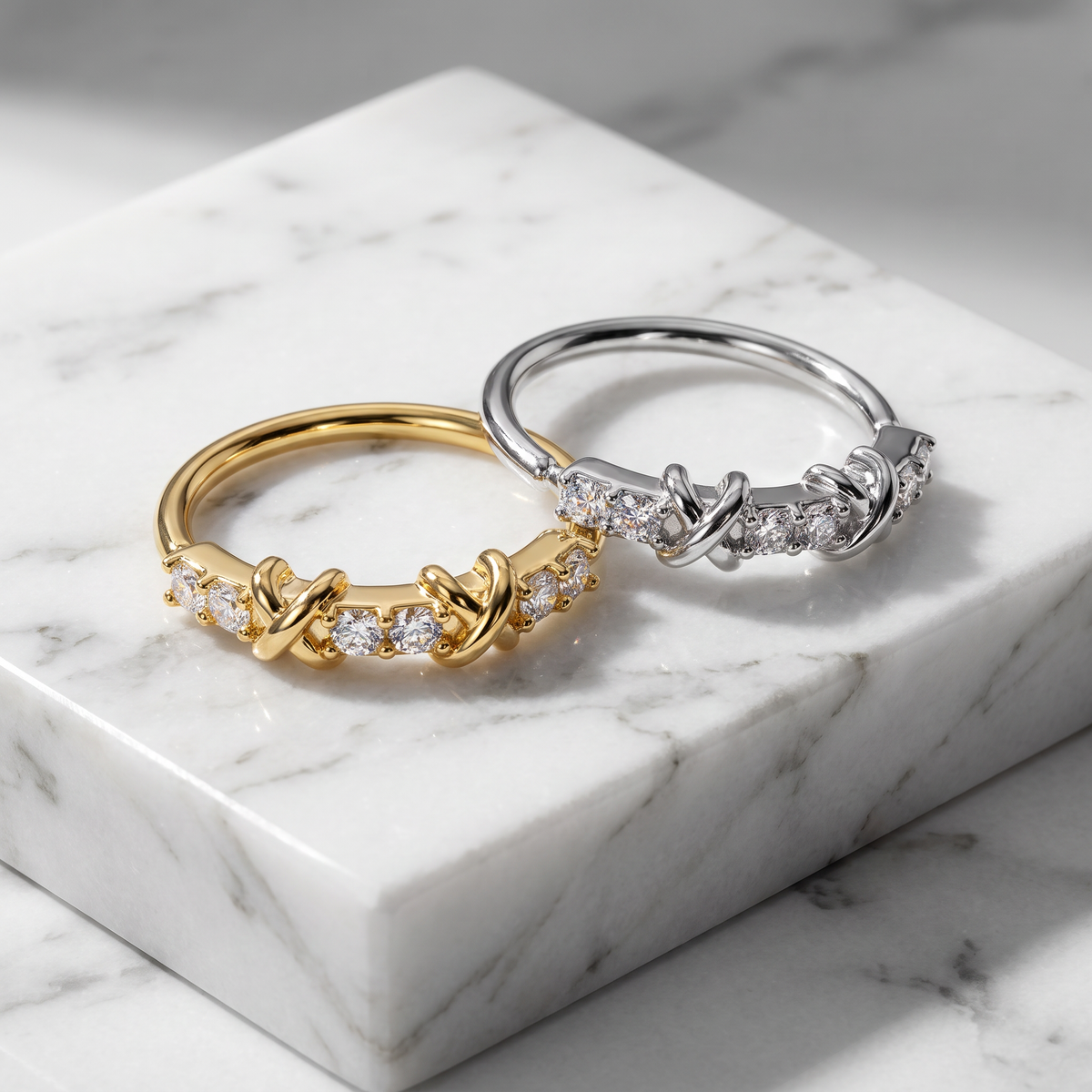 Two twisted knot rings with small clear stones, one in yellow gold and one in silver, displayed on a white marble block.