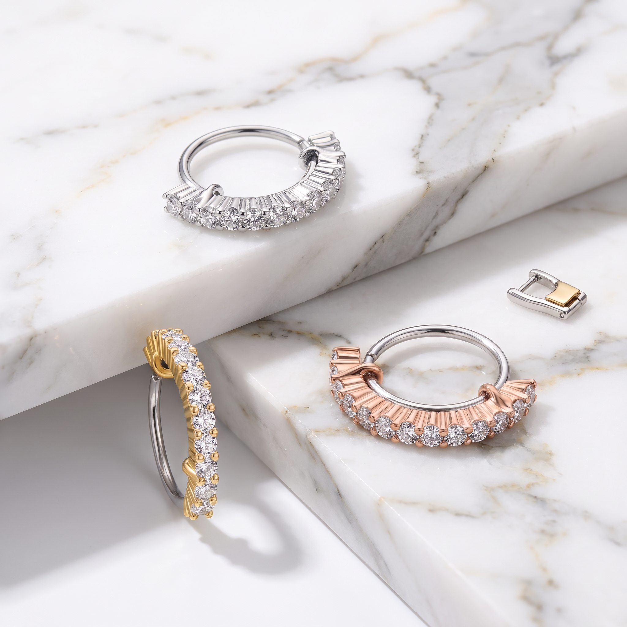 Three semi-hoop earrings displayed on a white marble surface: one silver with clear baguette stones, one rose gold with clear baguette stones, and one yellow gold with clear baguette stones. A small matching metal clasp is shown separately on the right.