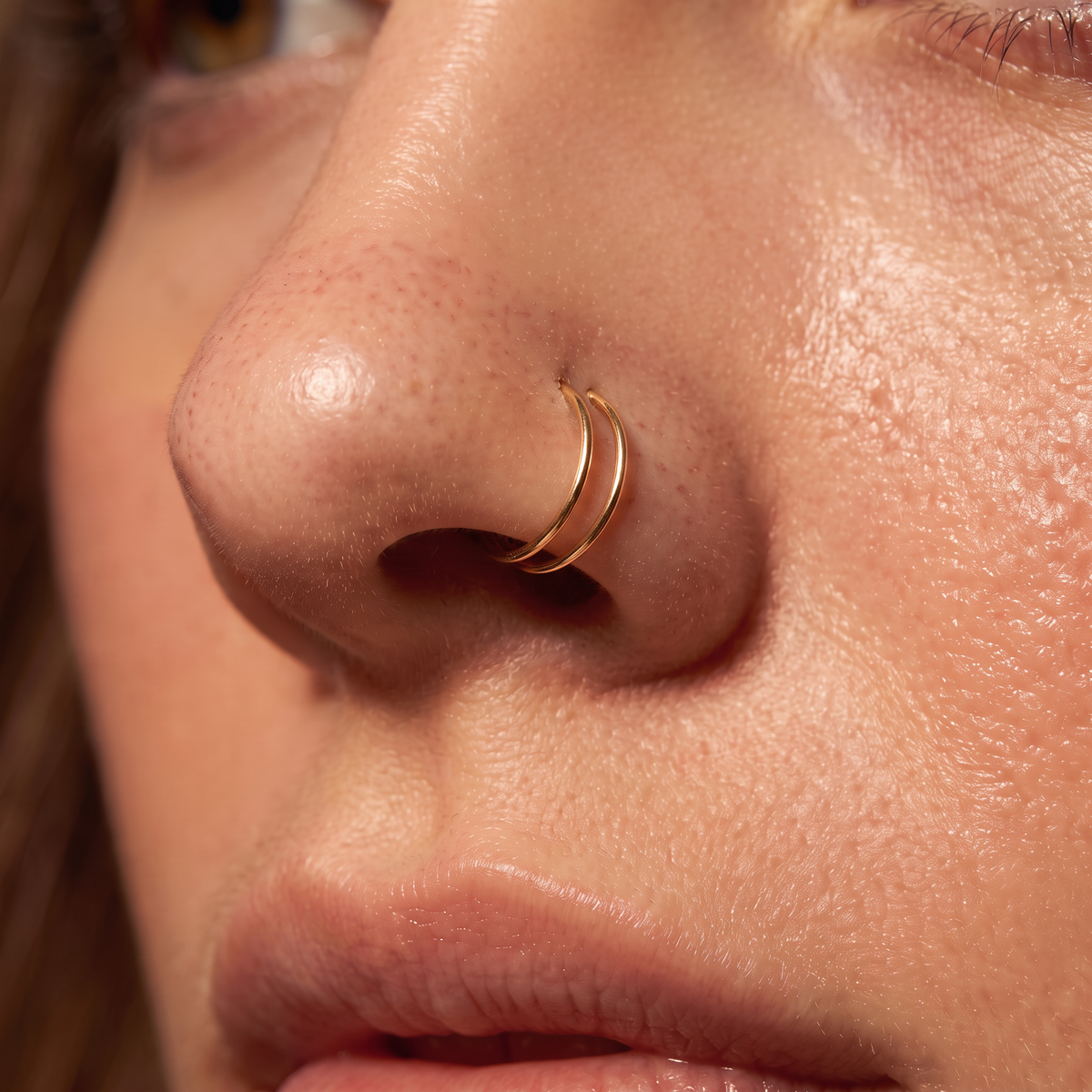 A close-up of a person's nose wearing a delicate double-band gold nose ring.