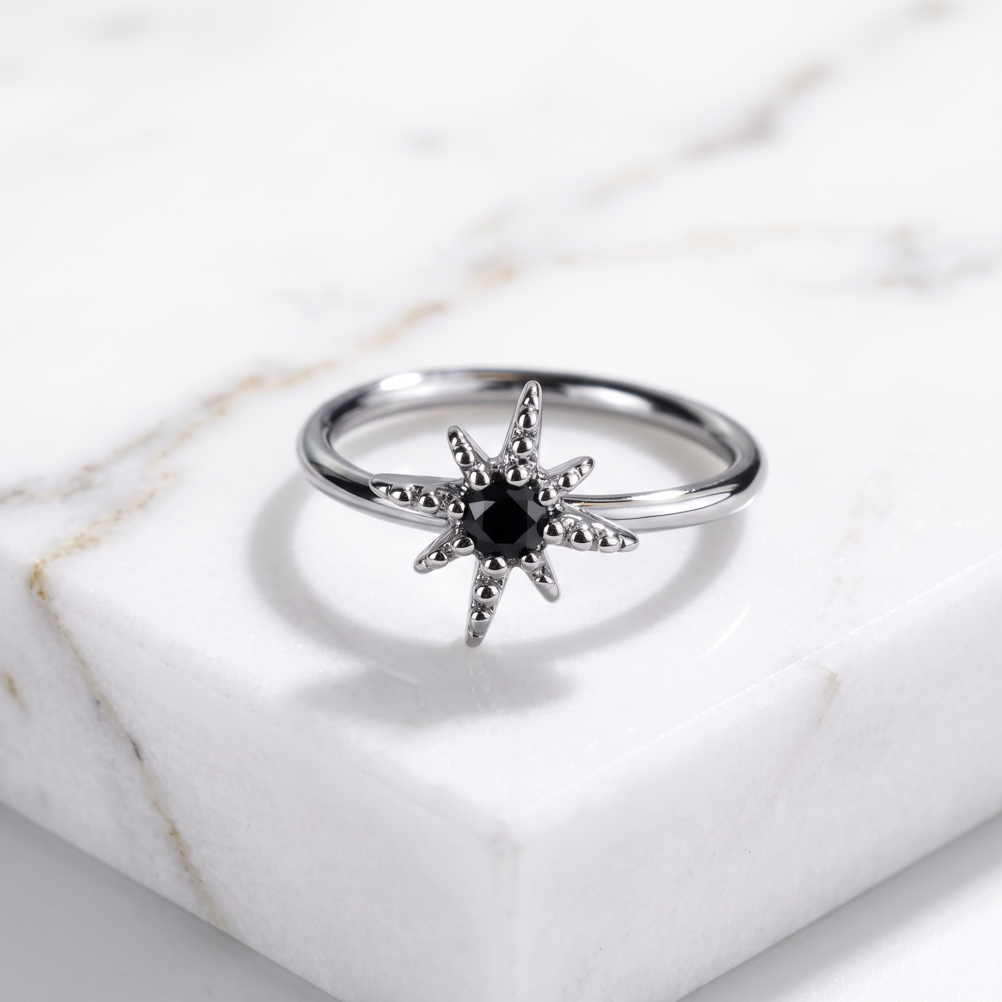 A silver ring shaped like a starburst, featuring a central black gemstone surrounded by small clear stones, displayed on a white marble block.