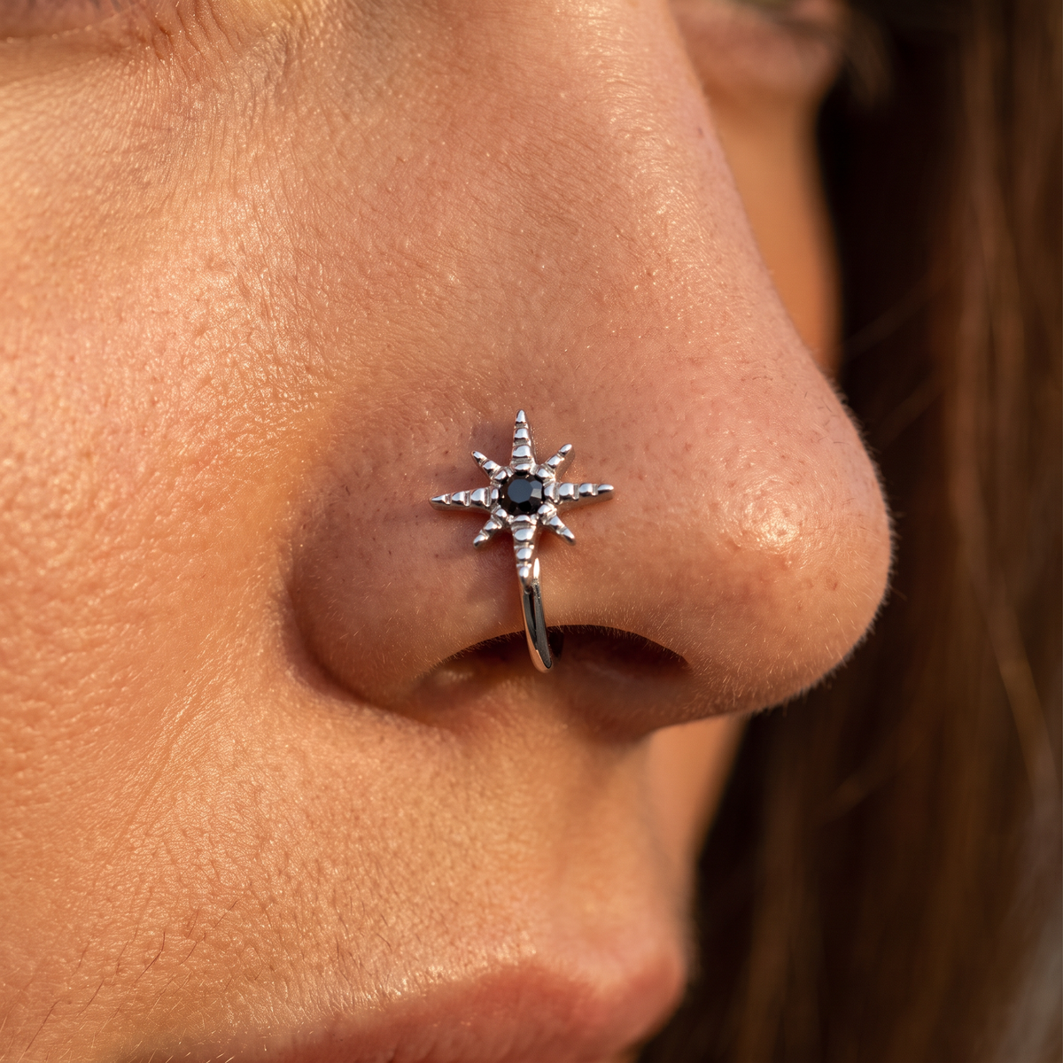 A close-up of a person's nose wearing a silver-toned starburst nose ring with a dark center stone and small clear stones along the points.