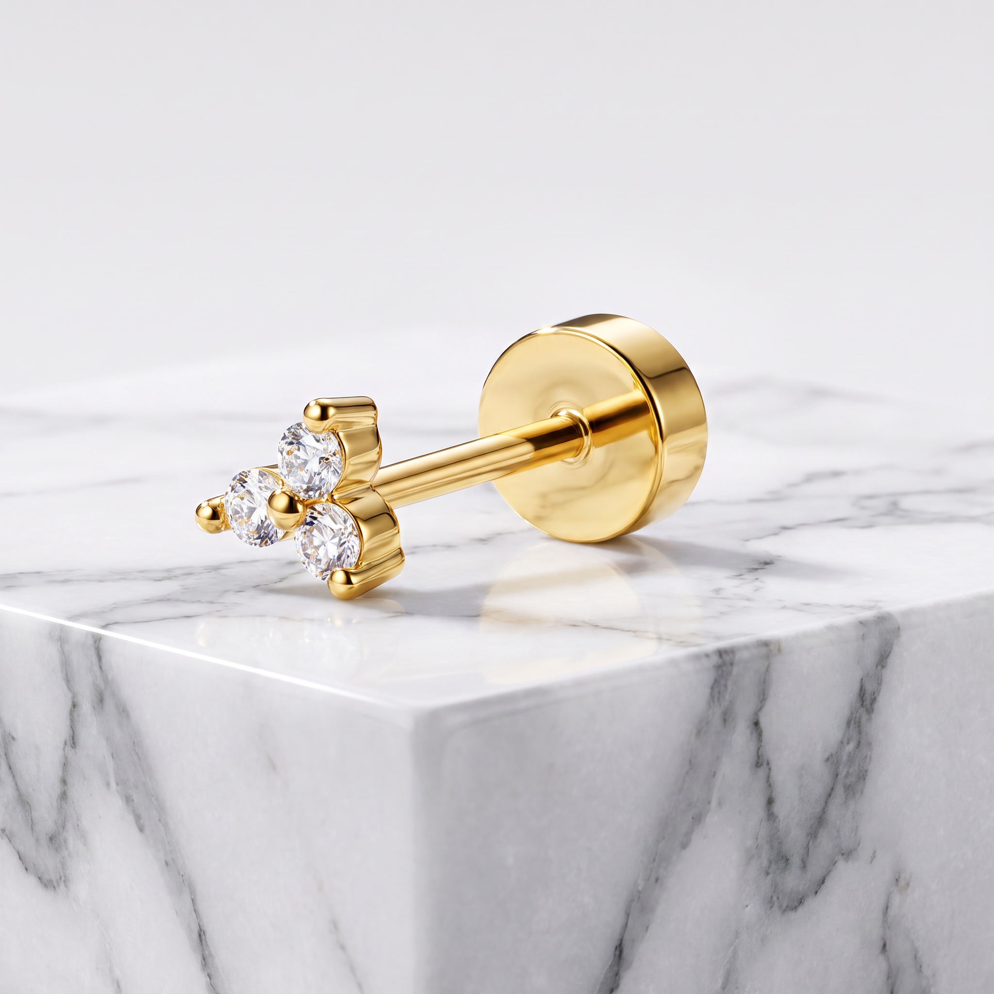 A gold post piercing stud with three clear gemstones, displayed on a white and gray marble block against a light gray background.