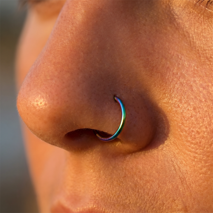 A close-up of a person's nose wearing a small curved nose ring with an iridescent rainbow finish.