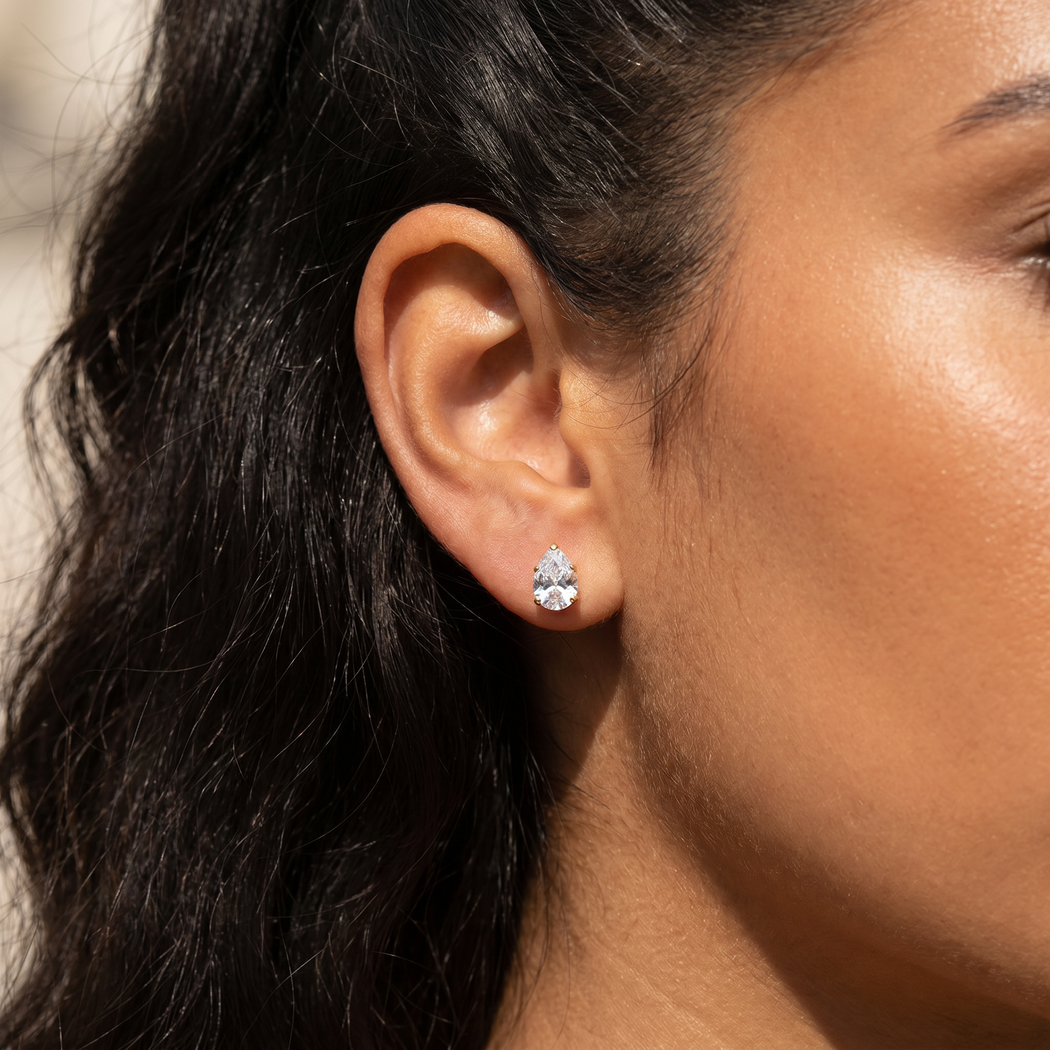 A close-up of a woman's ear wearing a small silver stud earring with a clear teardrop-shaped gemstone.