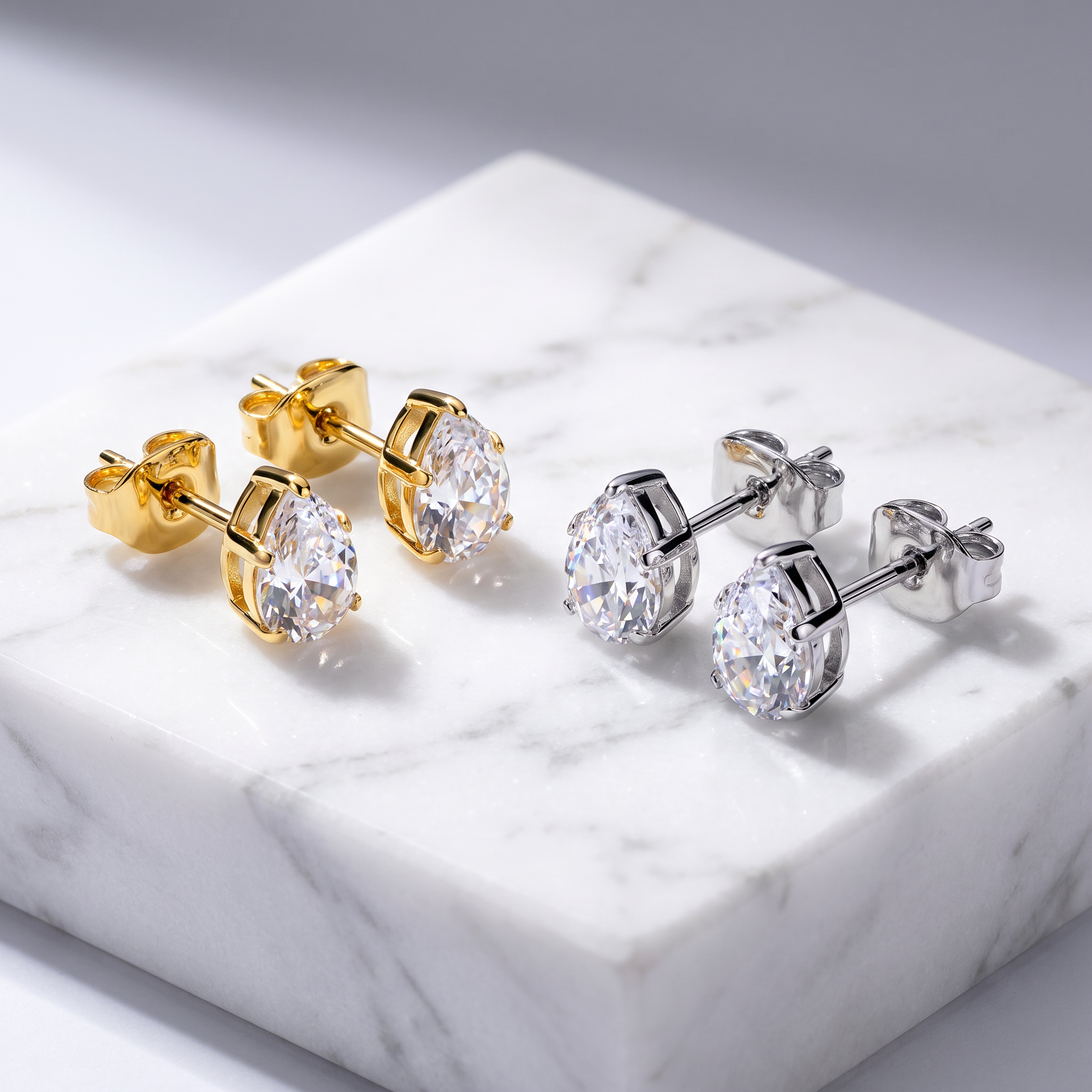Two pairs of oval stud earrings displayed on a white marble block. One pair is yellow gold with clear stones, and the other is silver with clear stones.