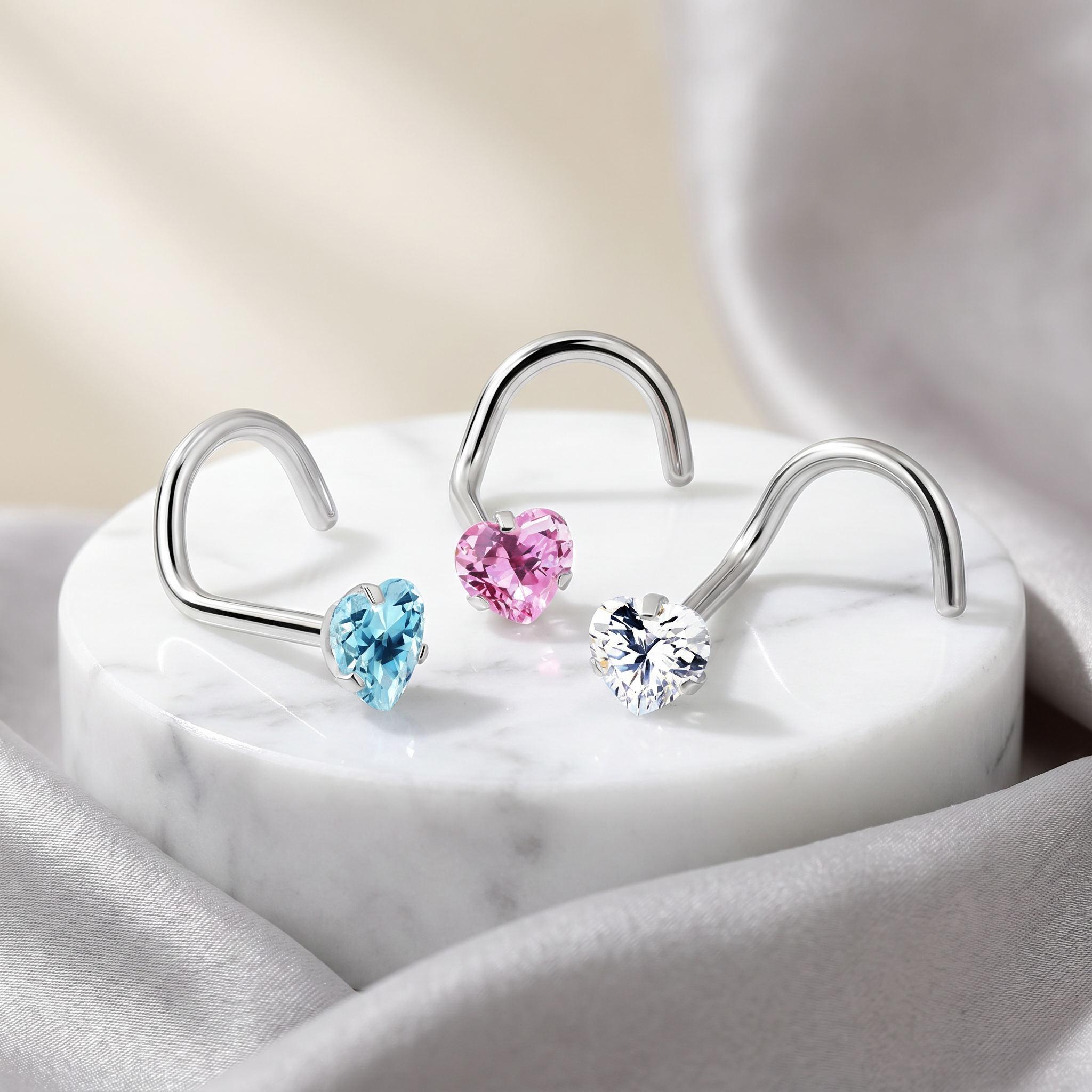 Three silver-toned U-shaped hoop earrings with heart-shaped crystal studs in light blue, pink, and clear, displayed on a white marble pedestal with soft gray fabric in the background.