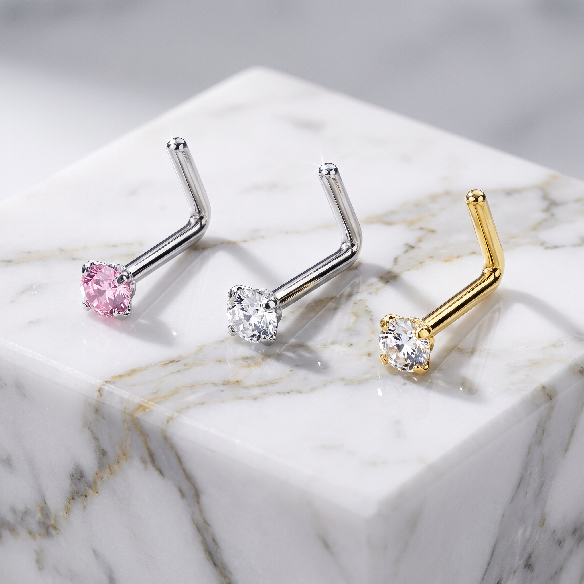 Three curved barbell-style stud earrings displayed on a white marble block. The left earring has a pink gemstone with a silver-toned post, the center earring has a clear round gemstone with a silver-toned post, and the right earring has a clear round gemstone with a gold-toned post.