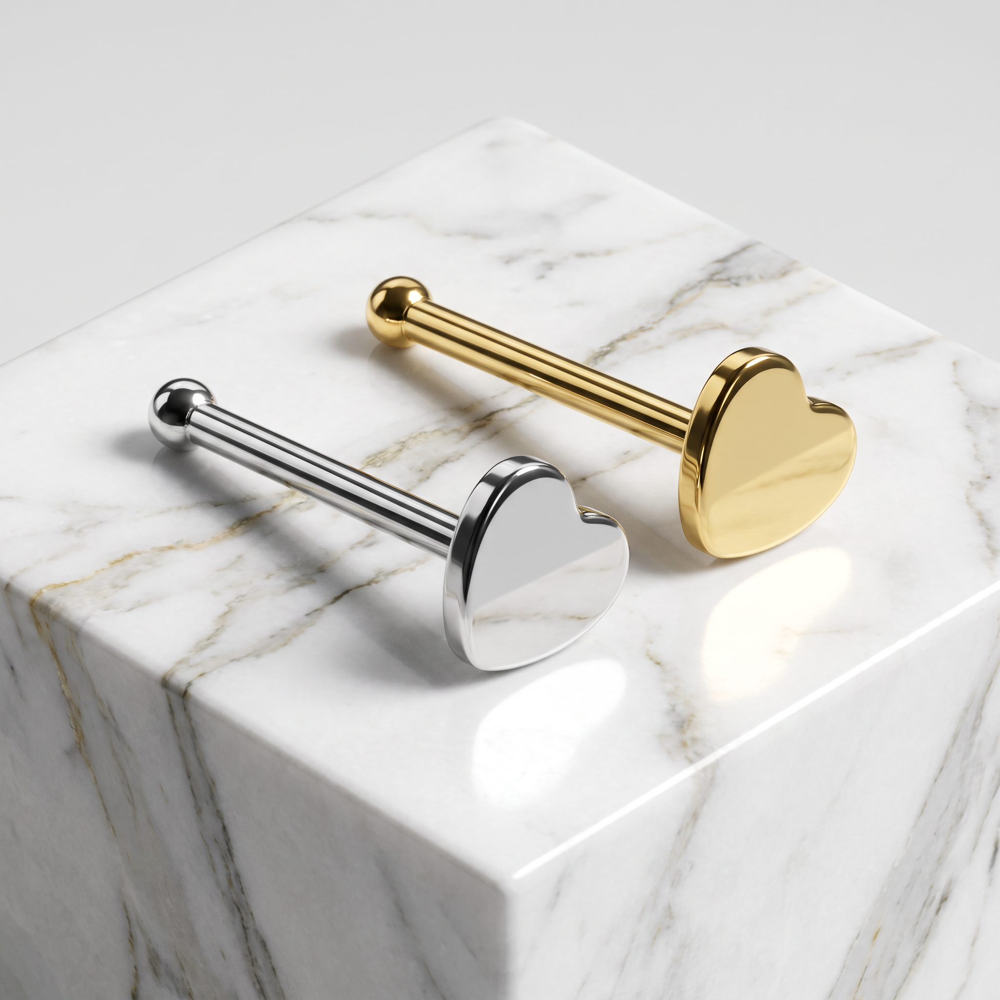 Two heart-shaped labret studs, one in polished silver and one in polished gold, displayed on a white marble cube against a light gray background.