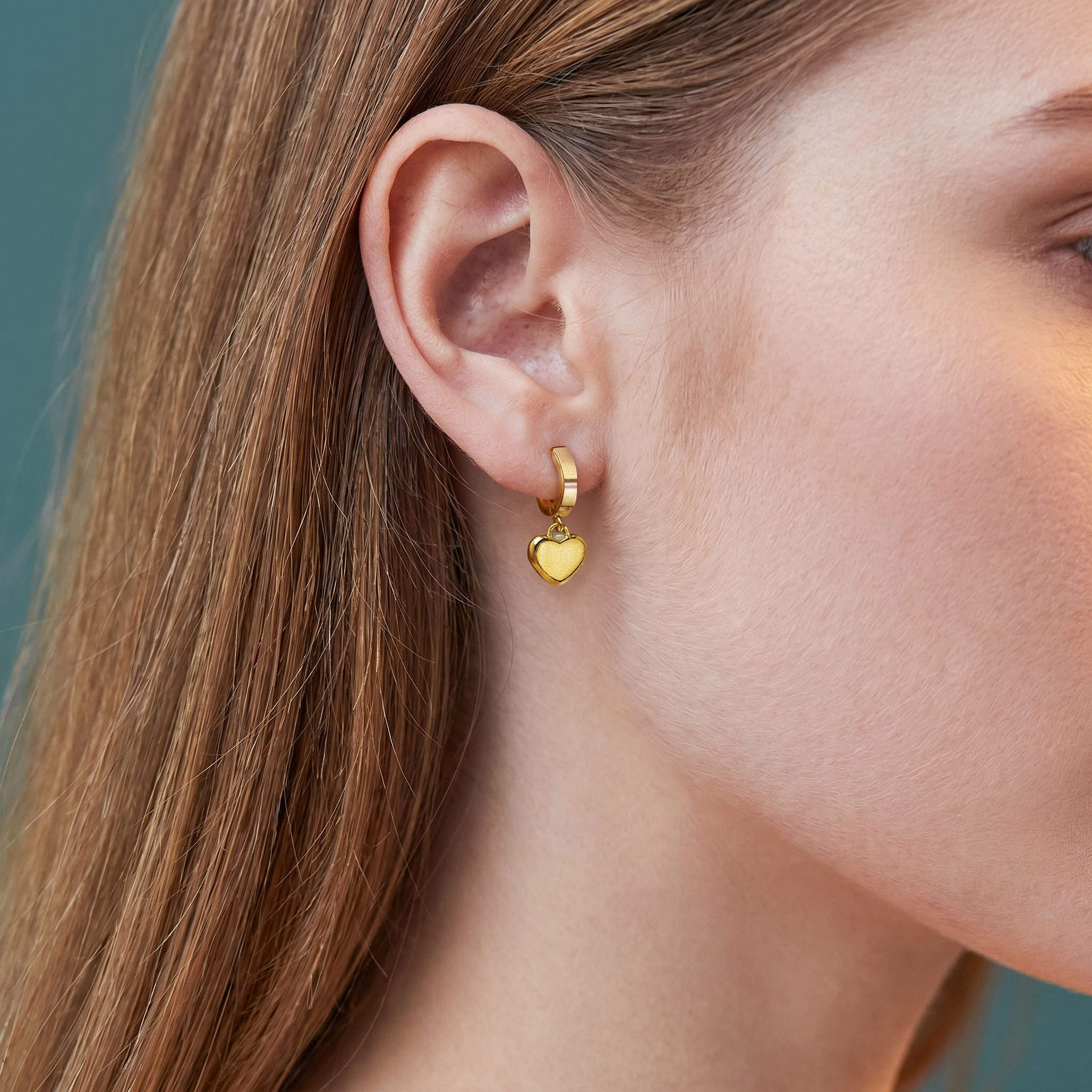 A close-up side profile of a woman wearing a small gold hoop earring with a dangling polished gold heart charm, set against a muted teal background.