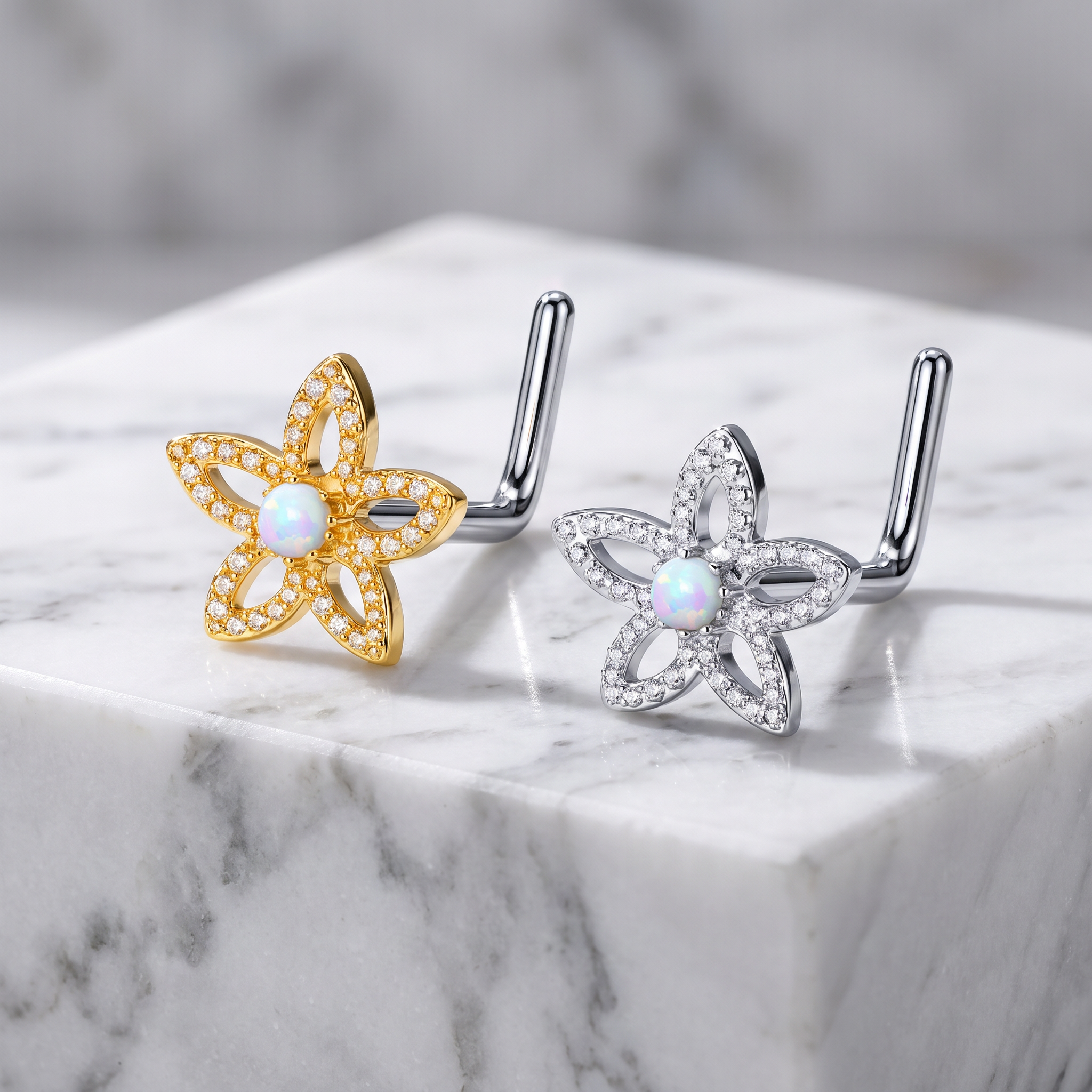 Two flower-shaped body jewelry piercings displayed on a white marble block. One is gold-toned with a central iridescent opal-like stone and small clear stones along the petals, and the other is silver-toned with the same center and petal detailing.