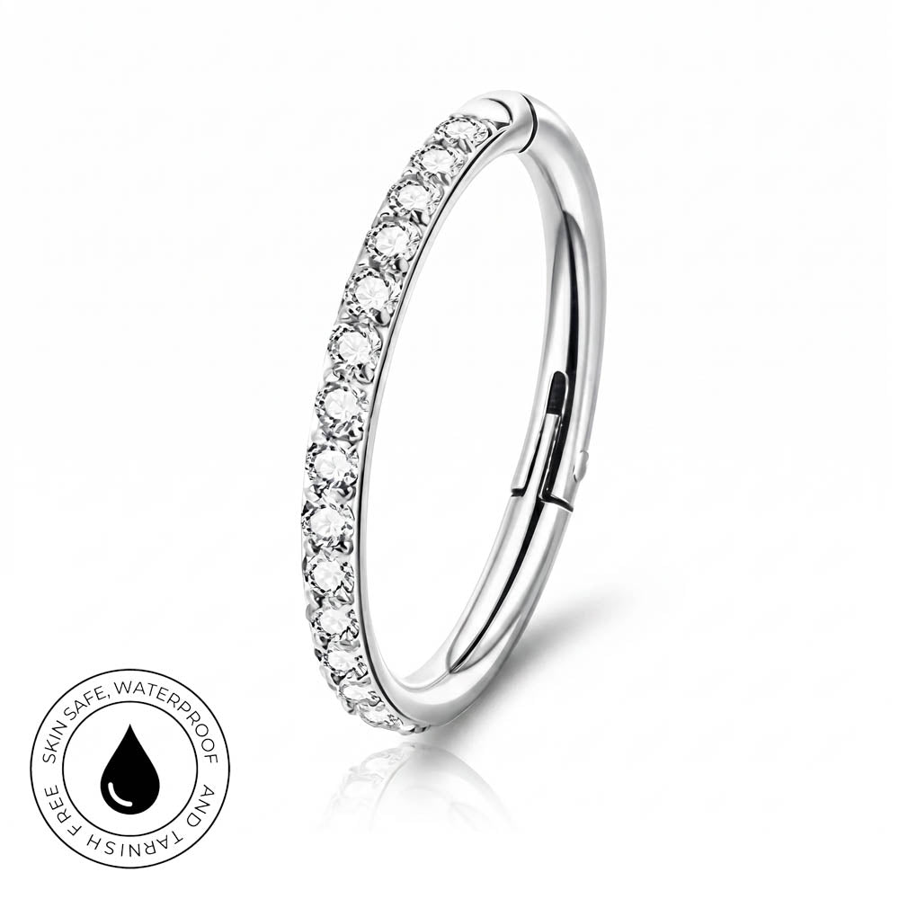 A silver-toned curved ring with a row of clear stones along the front, shown on a white background with a small circular skin-safe waterproof badge in the lower left.