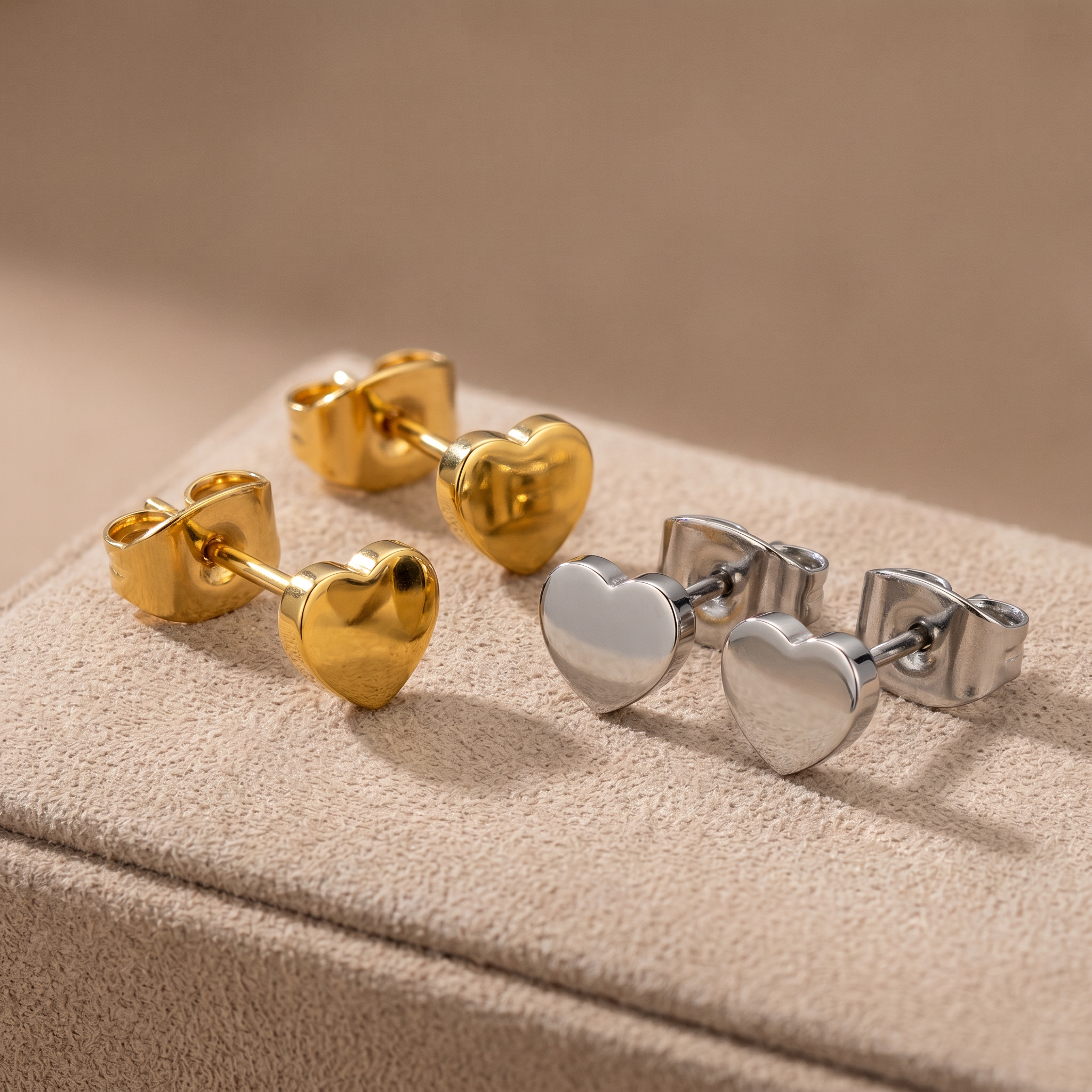 Two pairs of heart-shaped stud earrings displayed on a beige textured jewelry box. One pair is polished yellow gold, and the other is polished silver.
