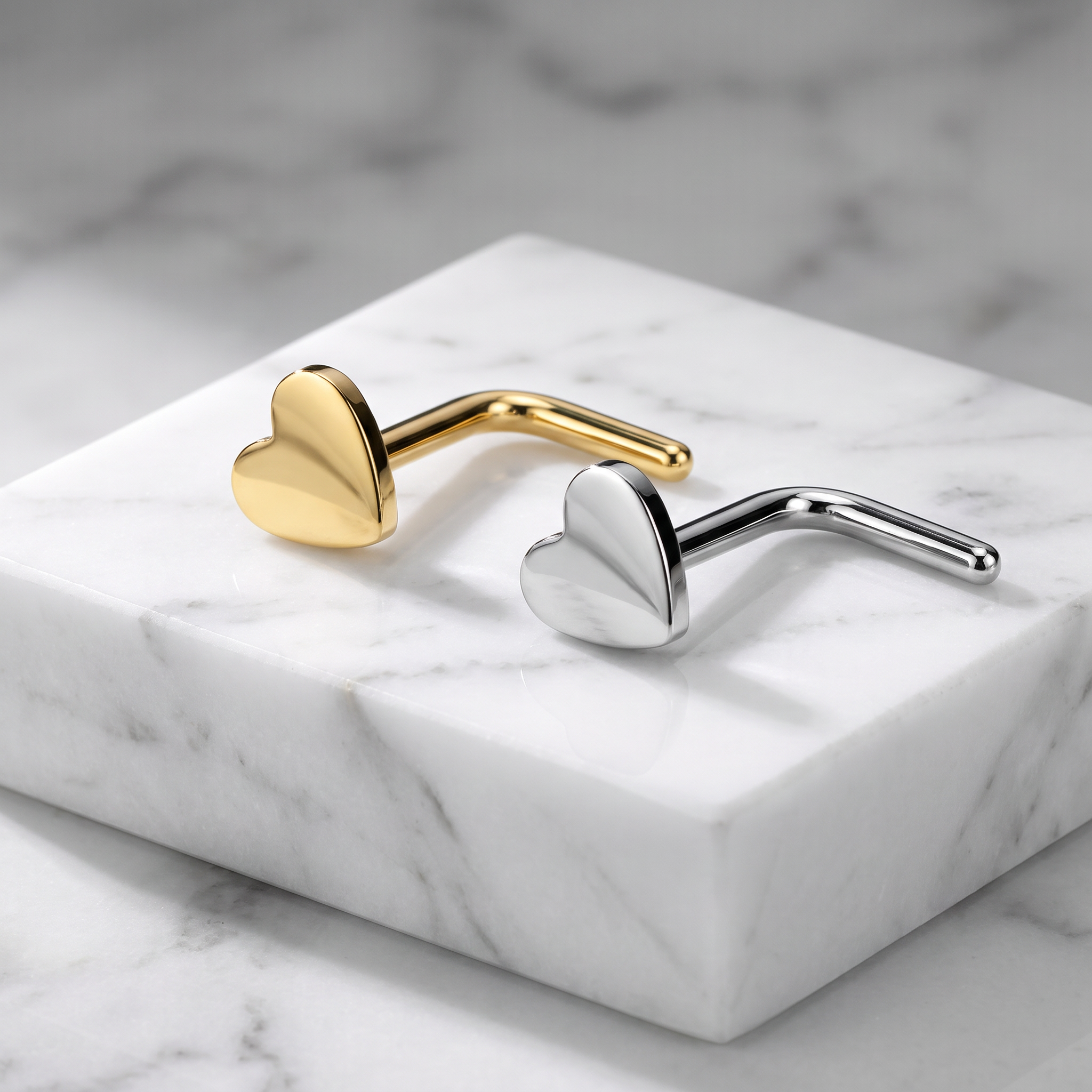 Two heart-shaped metal ear cuffs, one in polished gold and one in polished silver, displayed on a white marble block against a marble surface.