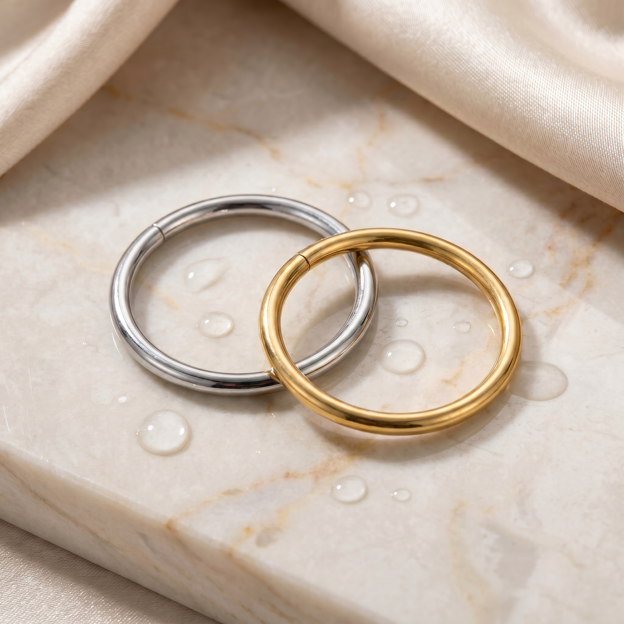 Two minimalist circular hoop earrings, one in polished silver and one in polished yellow gold, resting on a beige marble surface with water droplets and draped satin fabric.