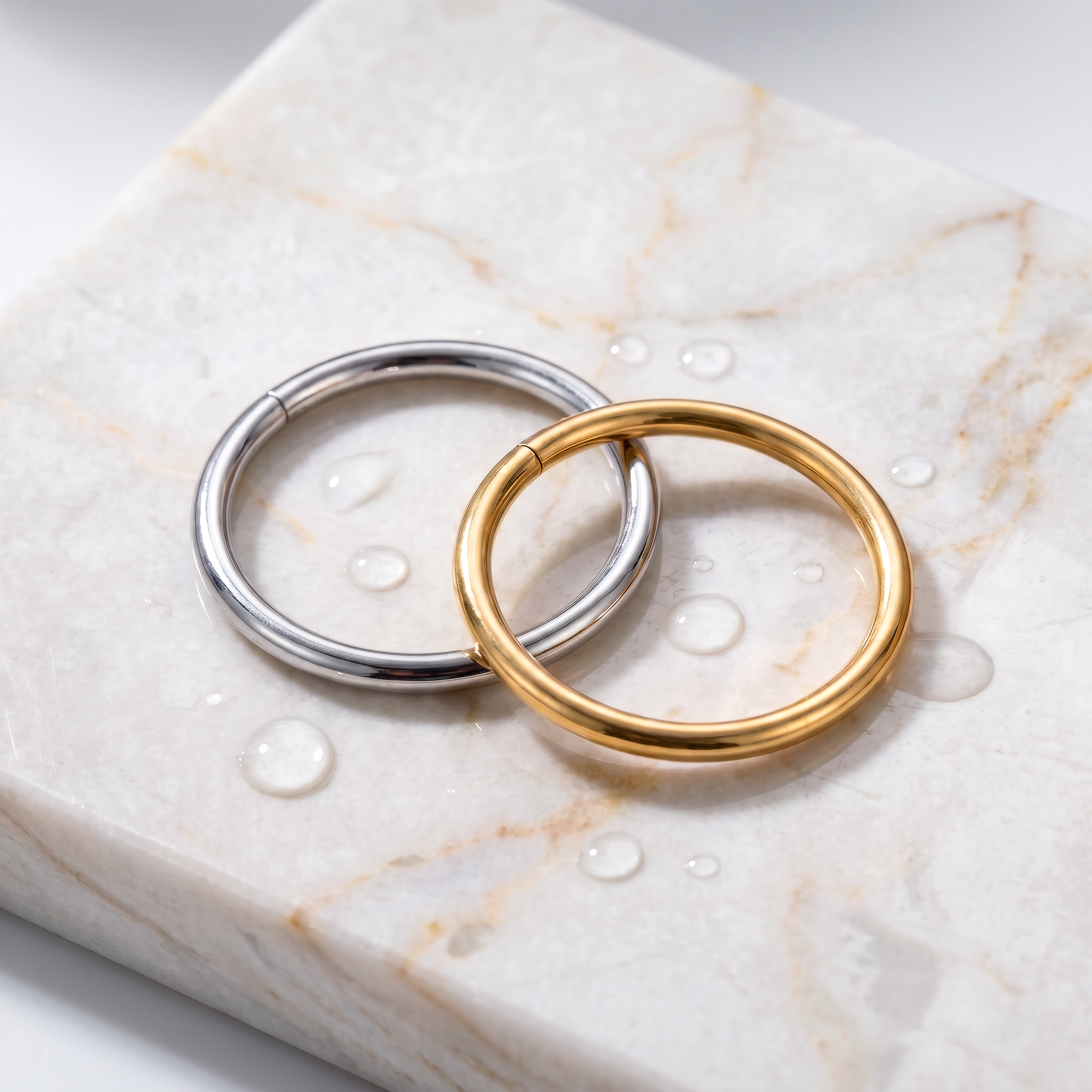 Two minimalist circular ring clips, one in polished silver and one in polished yellow gold, resting on a white marble slab with water droplets.