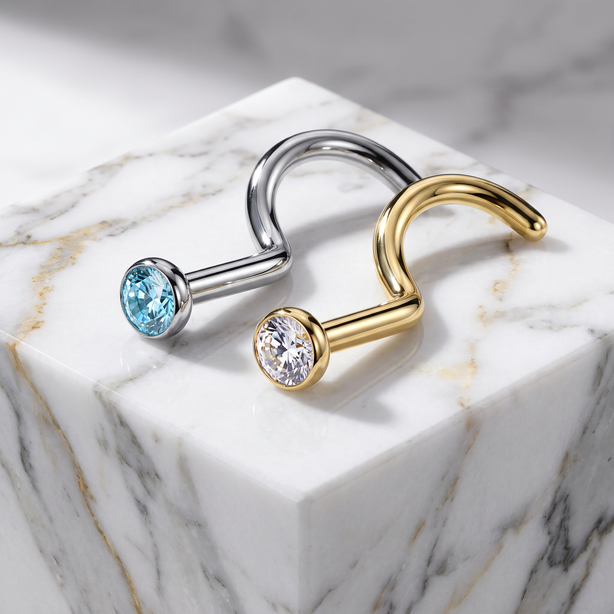 Two curved hoop-style earrings displayed on a white marble block. One earring is silver-toned with a light blue gemstone, and the other is gold-toned with a clear gemstone.
