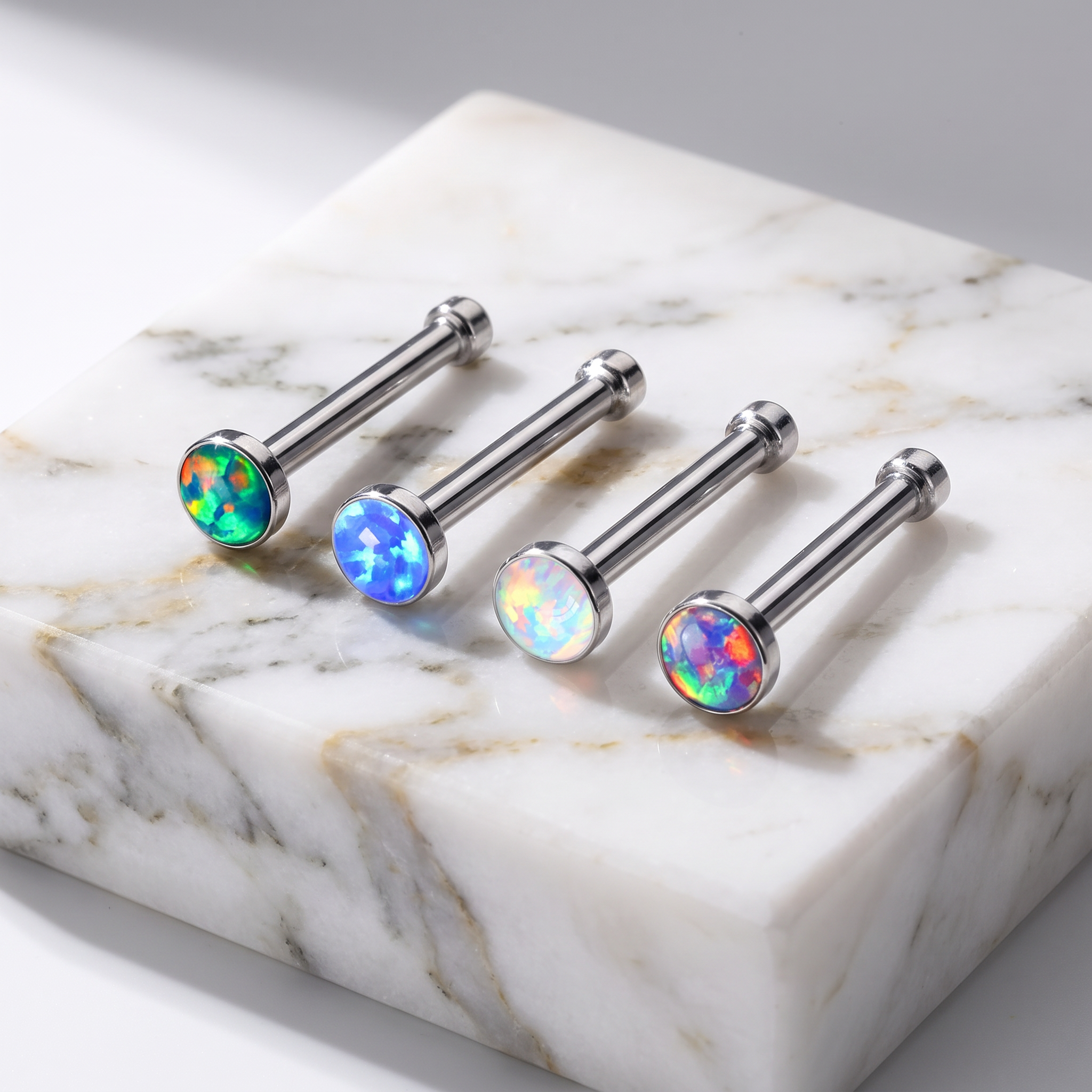 Four silver-toned labret ear piercings with round iridescent opal gemstone tops, arranged on a white marble block against a light gray background.