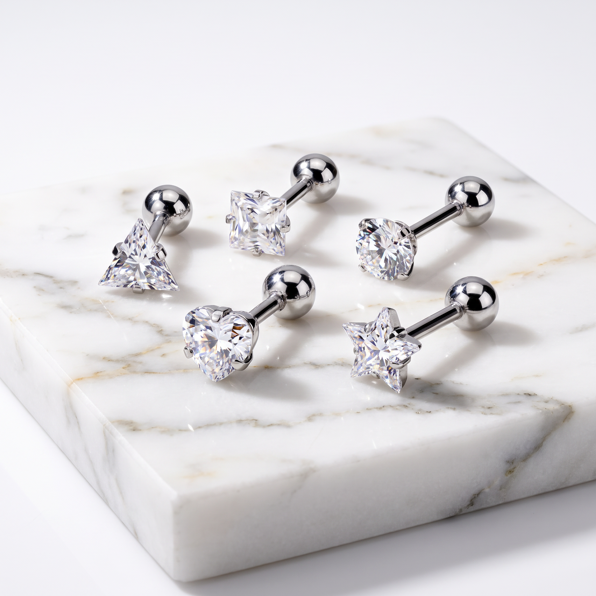 Five silver-toned labret ear piercings with clear faceted gemstones in different shapes, arranged on a white marble block against a white background.
