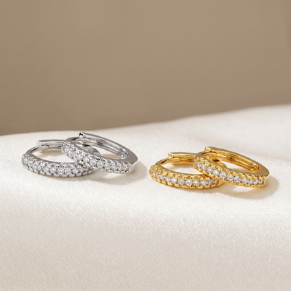 Two pairs of small hoop earrings with pavé-set clear stones, shown side by side on a soft cream fabric surface against a warm beige background. One pair is silver-toned and the other is yellow gold-toned.