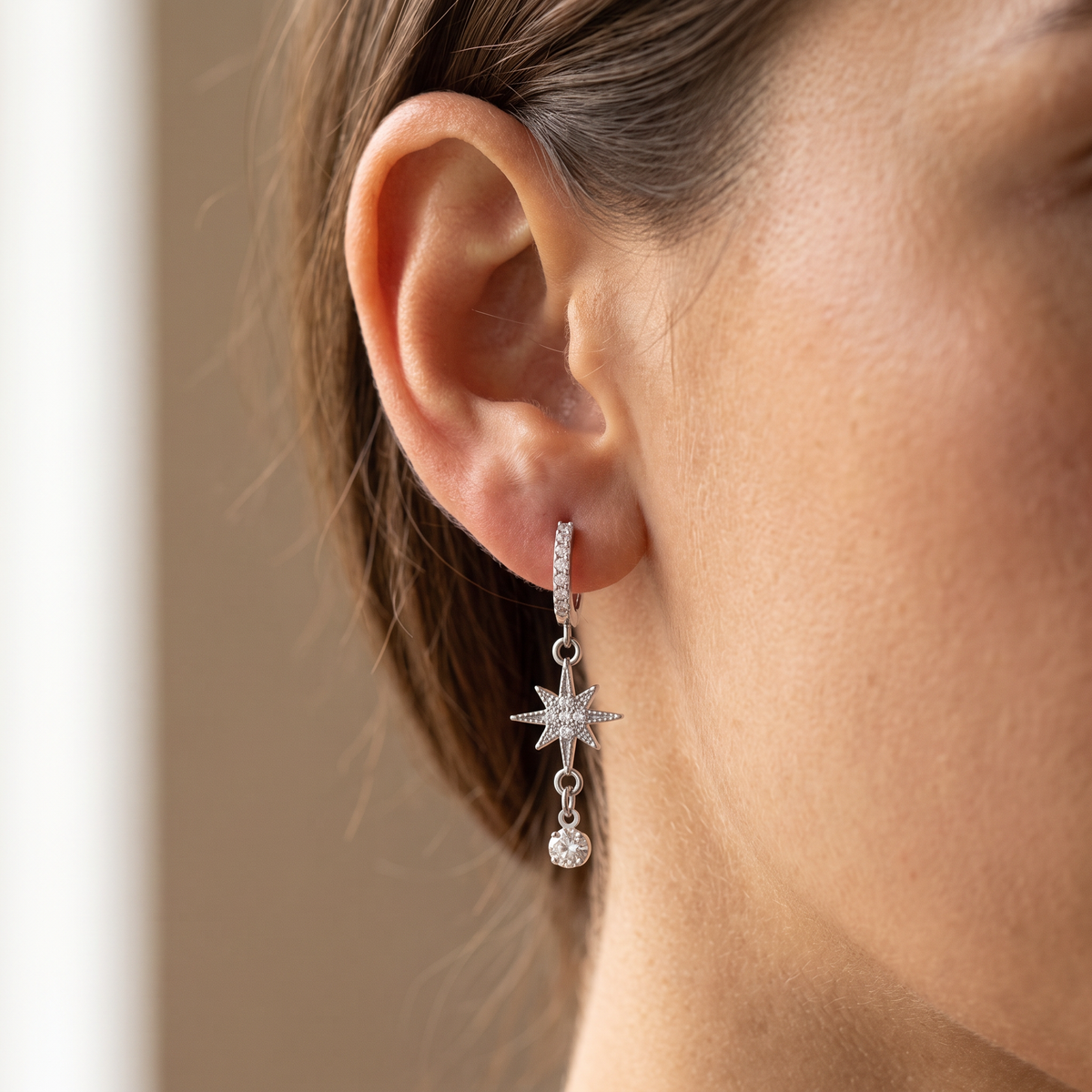 A close-up of a model's ear wearing a silver huggie hoop earring with a dangling starburst charm and a small clear crystal drop.