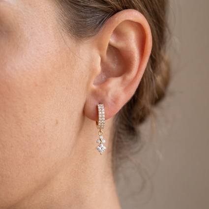 A close-up of a person's ear wearing a small gold huggie hoop earring adorned with clear pavé stones and a dangling clear star-shaped crystal.
