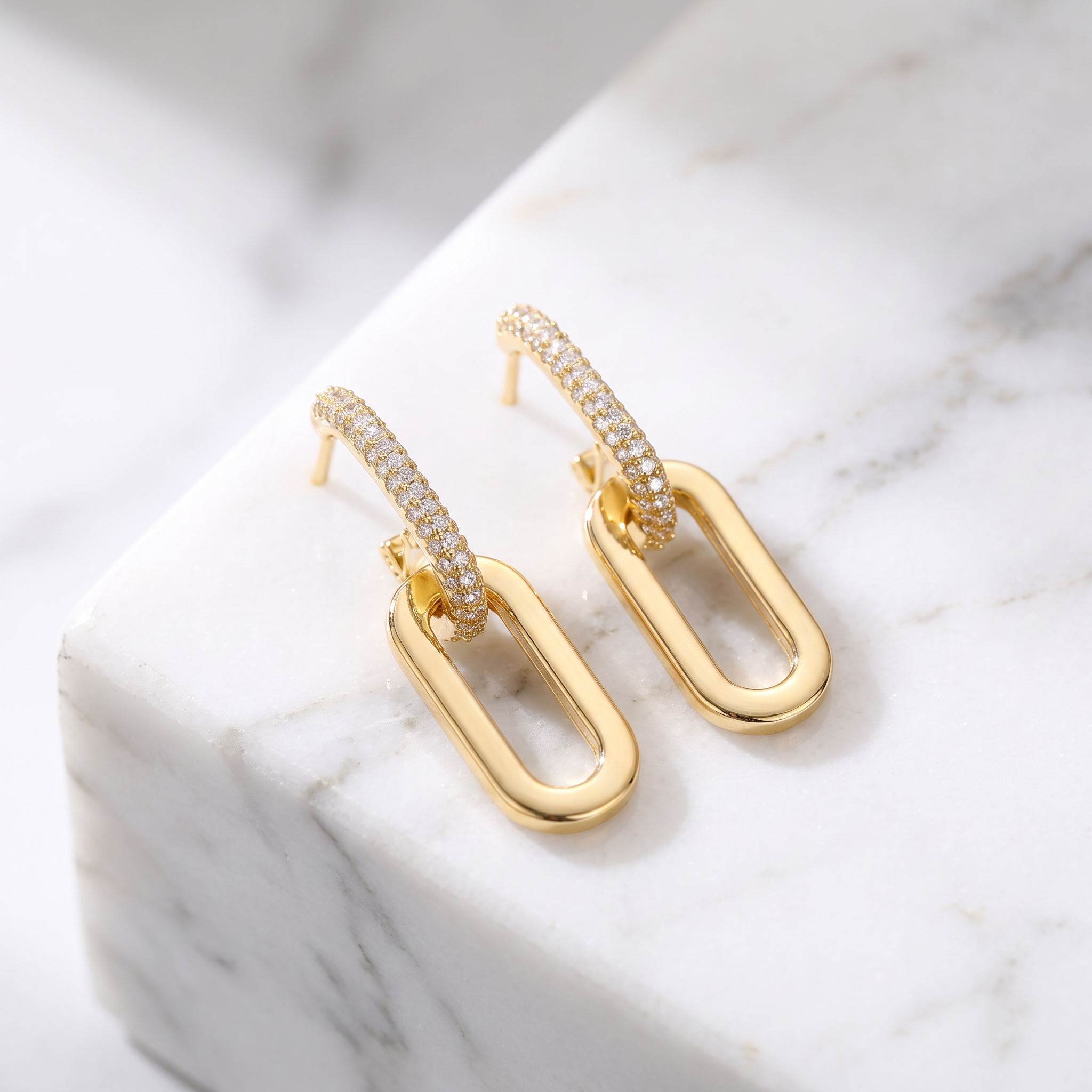 A pair of gold hoop earrings with pavé-set clear stones and elongated oval drop elements, displayed on a white marble block.