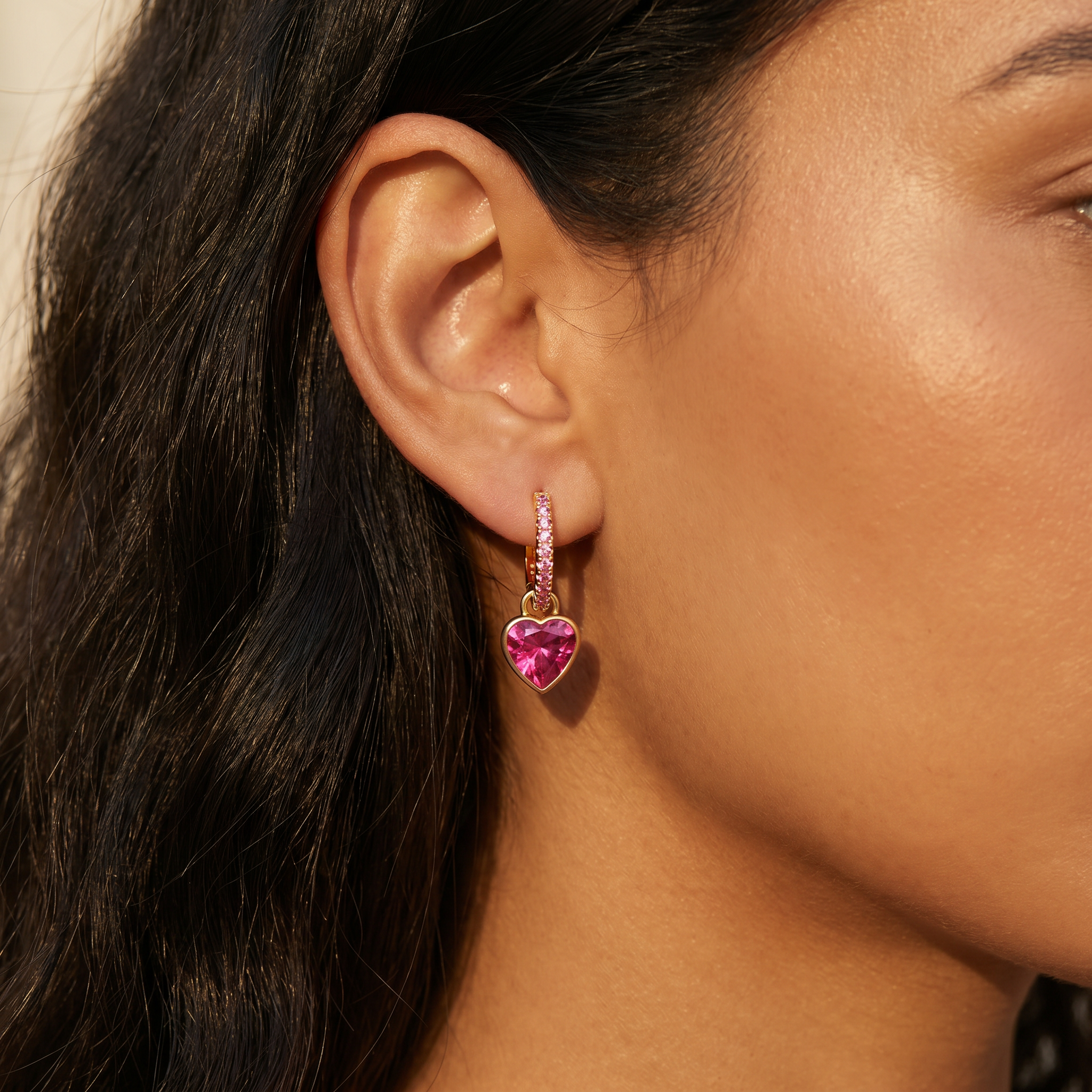 A close-up of a woman's ear wearing a gold huggie hoop earring with a dangling heart-shaped pink gemstone.