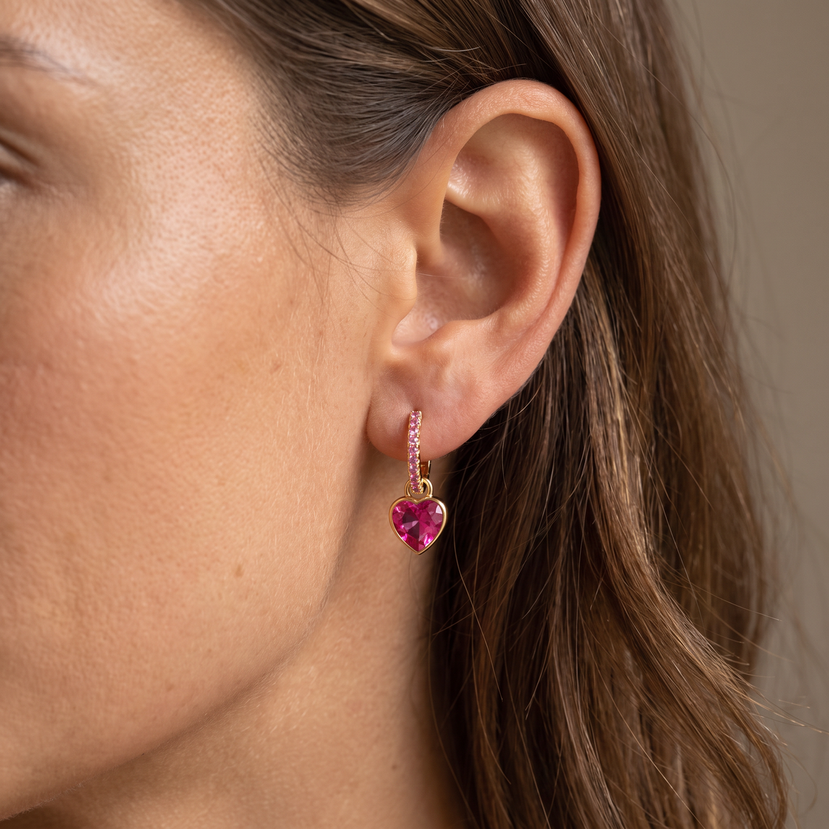 A close-up of a model's ear wearing a gold huggie hoop earring adorned with small pink stones and a dangling heart-shaped pink gemstone.