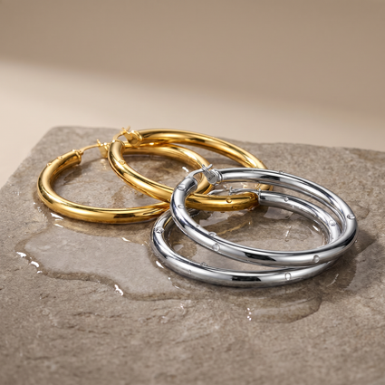 Two polished hoop earrings, one gold-toned and one silver-toned, resting on a wet stone surface against a warm beige background.