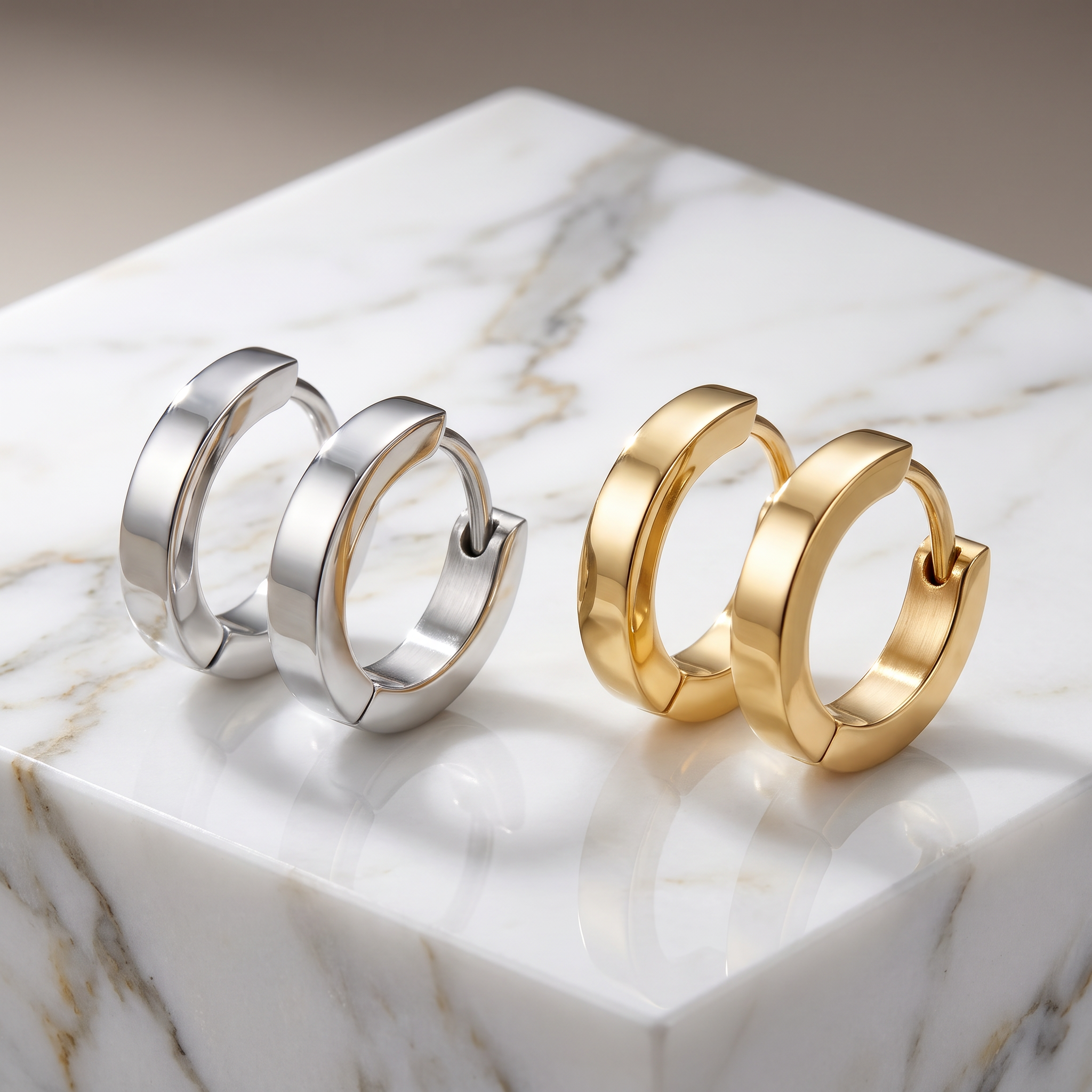 Two pairs of polished huggie hoop earrings, one in silver and one in yellow gold, displayed on a white marble block.