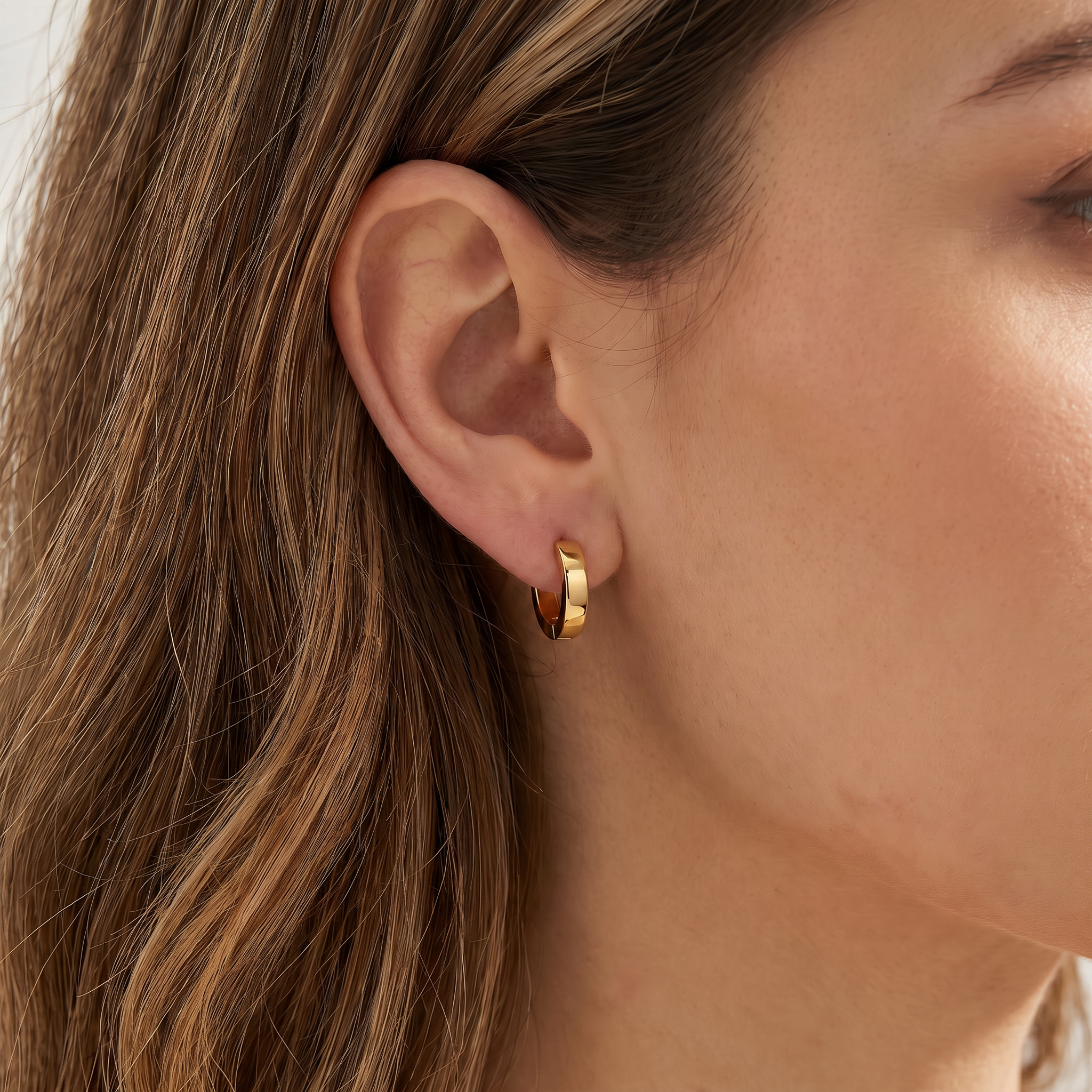 A close-up of a woman's ear wearing a small gold huggie hoop earring.
